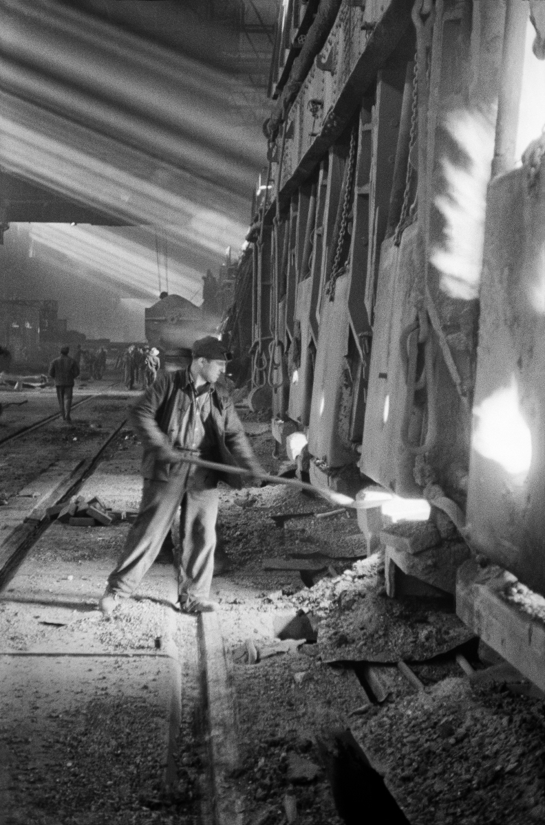 A man in work clothes uses a shovel to throw coke into a furnace hole in a huge structure.