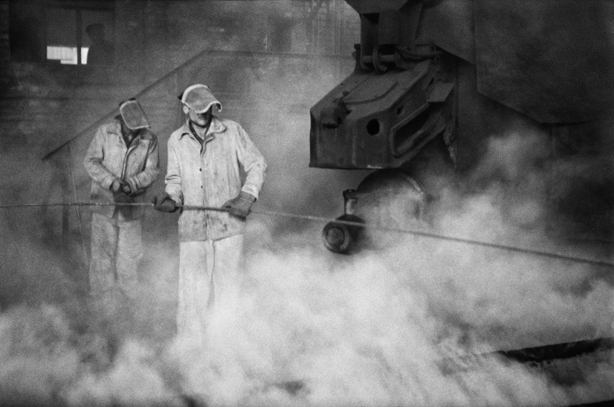 Two men in metallurgical clothes hammer a long rod into the foundations of a huge, steaming structure.