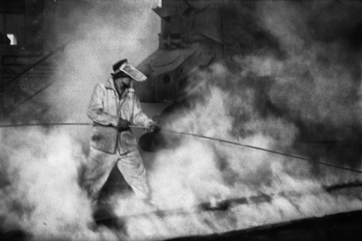 A man in a metallurgical costume drives a long rod into the foundations of a huge, steaming structure.