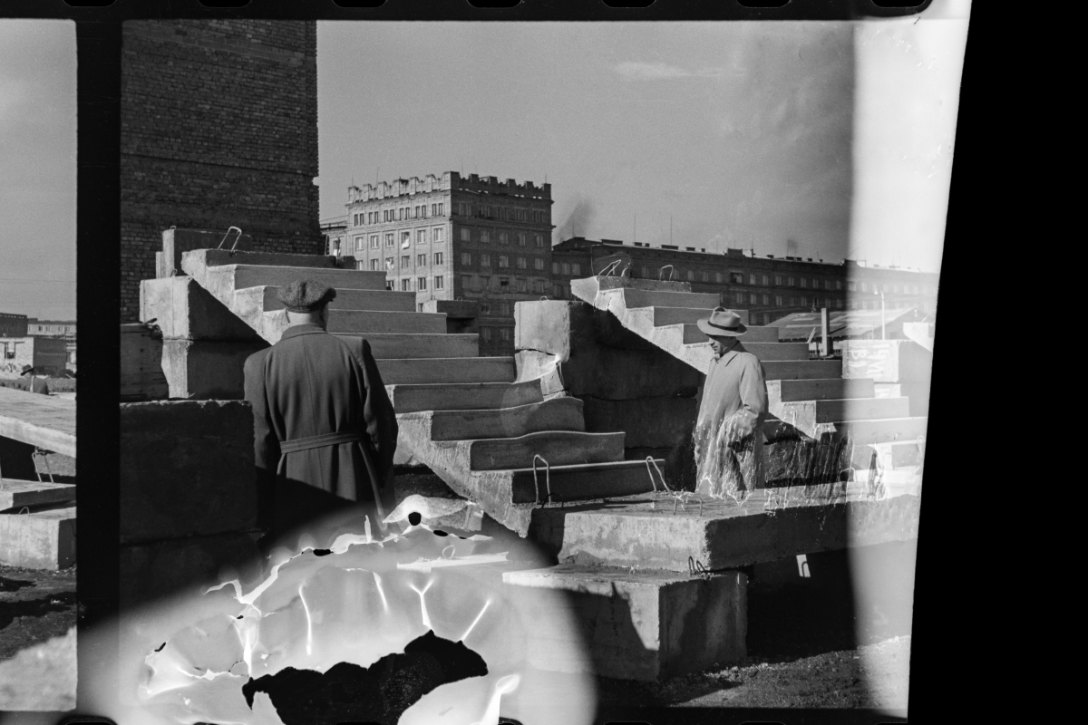 Men in coats and hats look at the display of prefabricated stairs. In the background, apartment blocks. Negative damaged while developing or burnt.