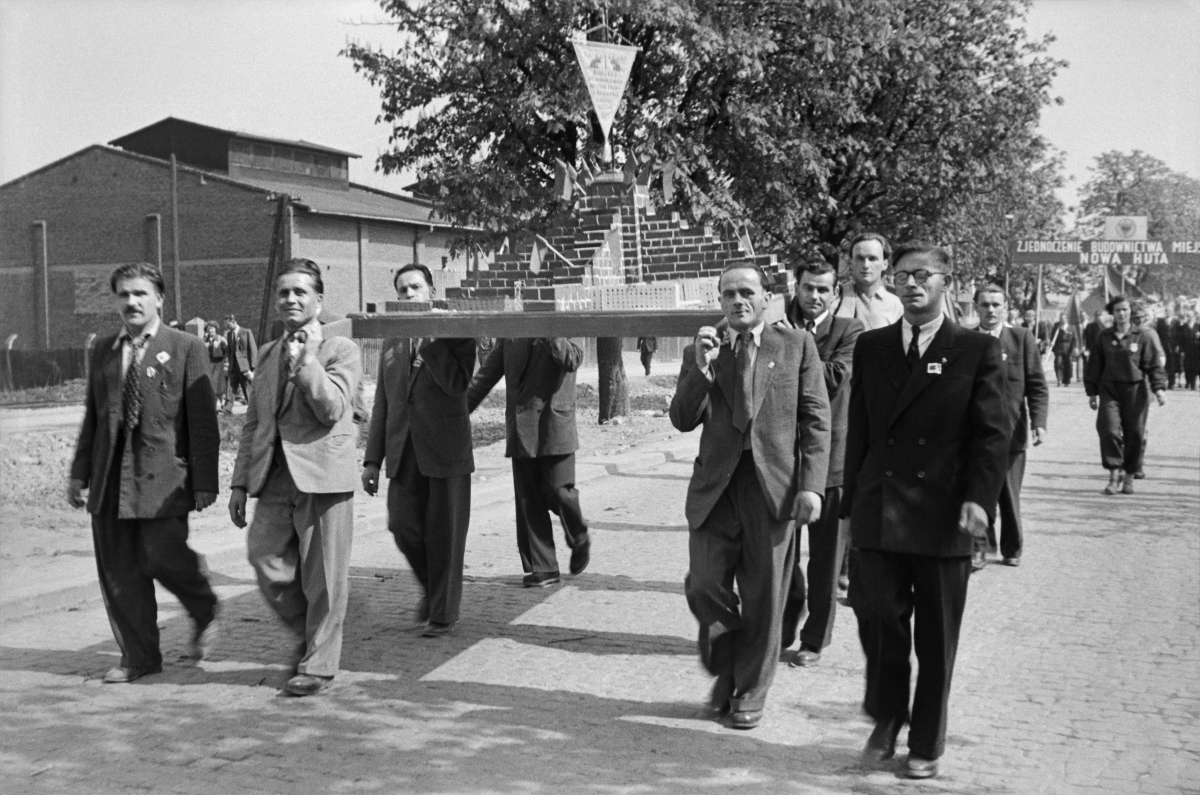 Men in suits march down the street, carrying a model of a pyramidal brick structure on their shoulders.