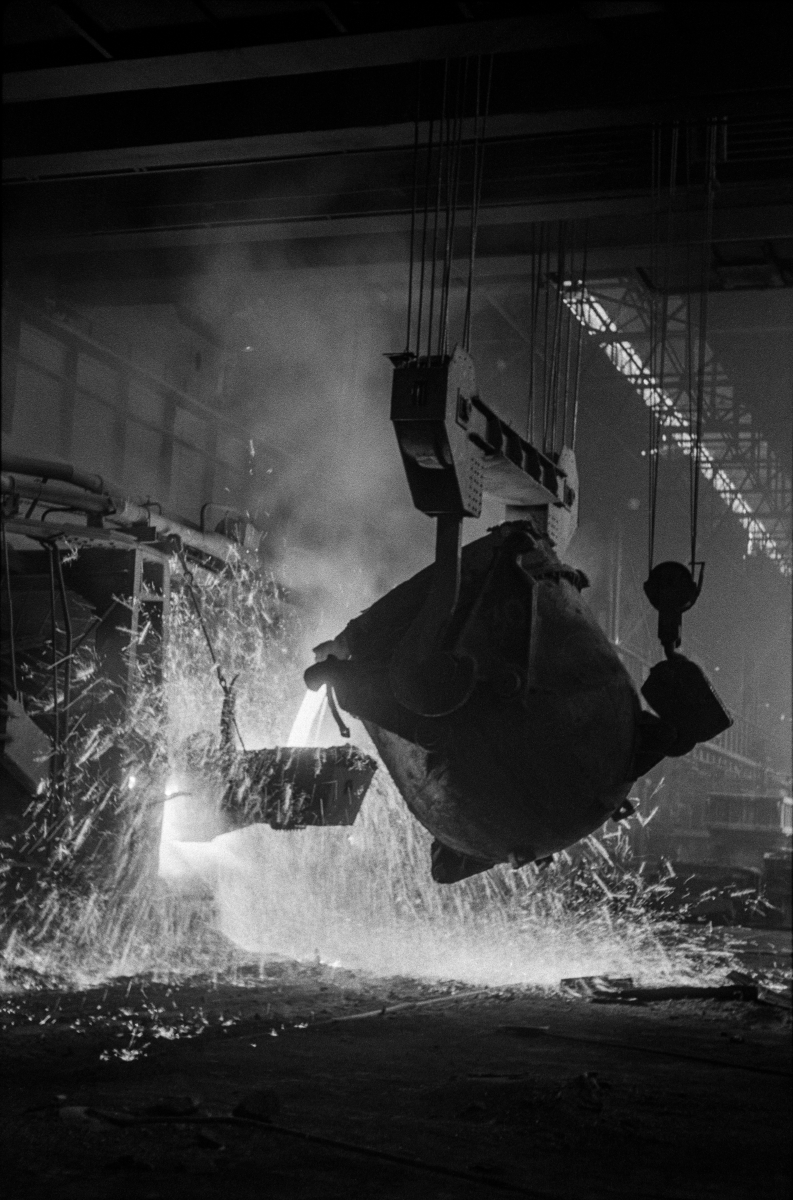 From the mighty, tilted vat, molten steel flows into a smaller, sparkling vessel.