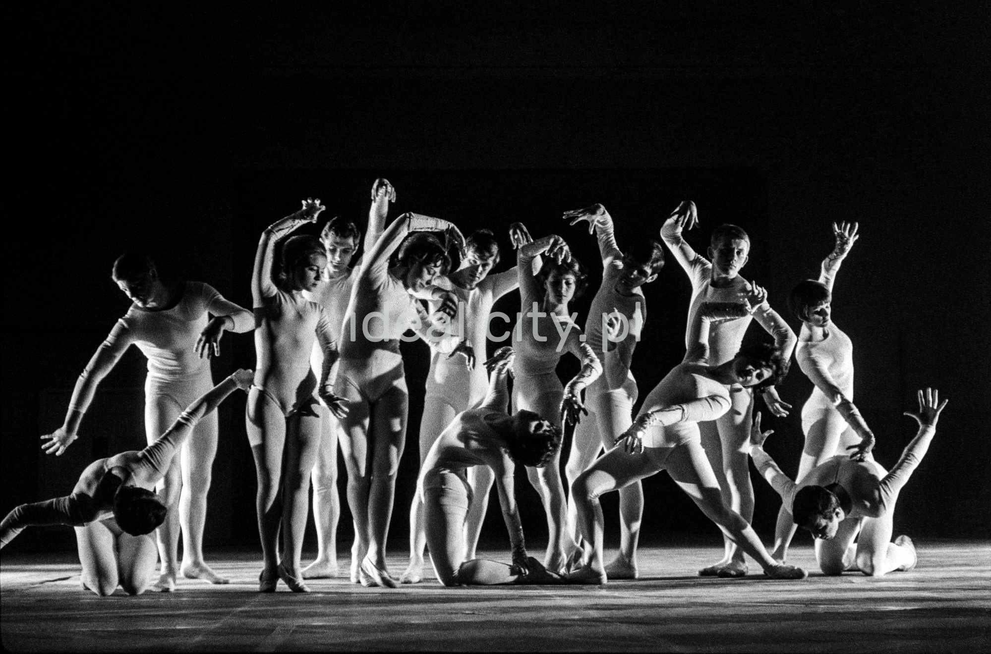 A group of dancers in tight-fitting costumes perform a collective figure on stage with a black background.