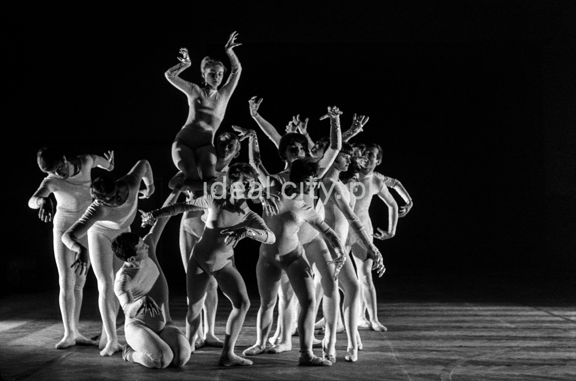 A group of dancers in tight-fitting costumes perform a collective figure on stage with a black background.