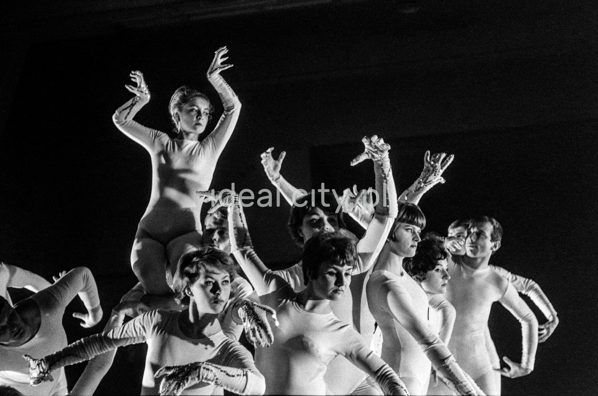 A group of dancers in tight-fitting costumes perform a collective figure on stage with a black background.