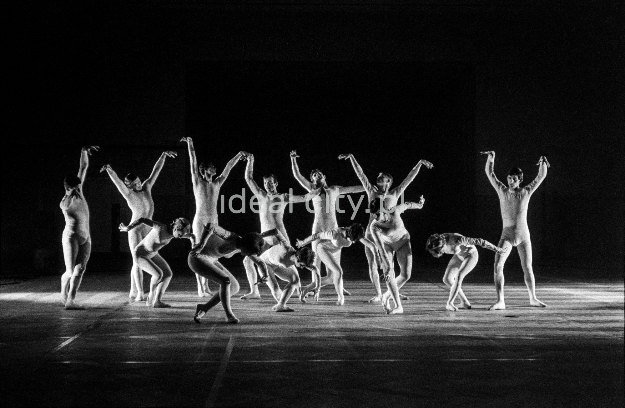 A group of dancers in tight-fitting costumes perform a collective figure on stage with a black background.