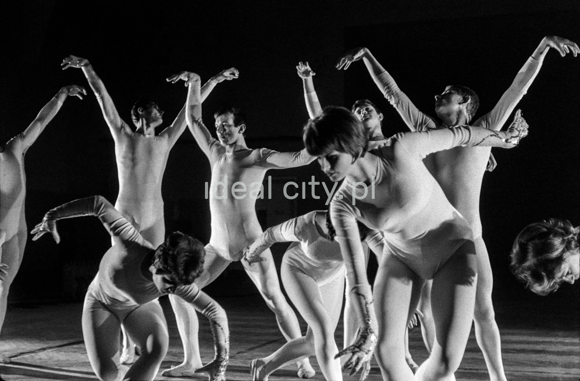 A group of dancers in tight-fitting costumes perform a collective figure on stage with a black background.