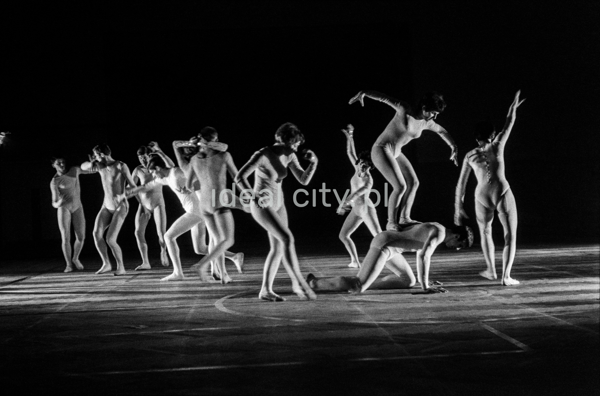 A group of dancers in tight-fitting costumes perform a collective figure on stage with a black background.