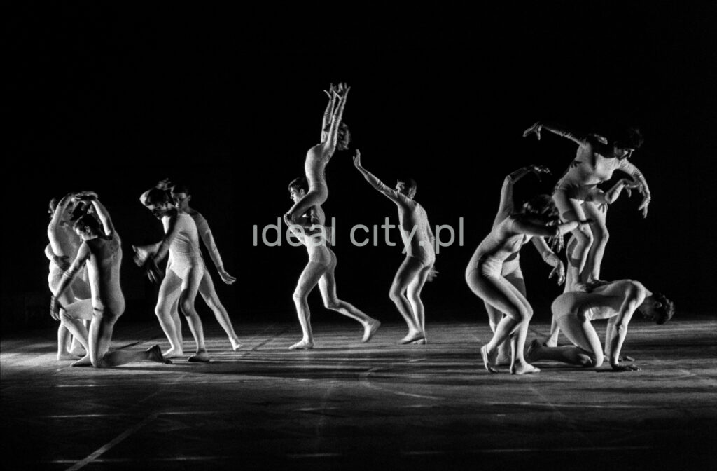 A group of dancers in tight-fitting costumes perform a collective figure on stage with a black background.