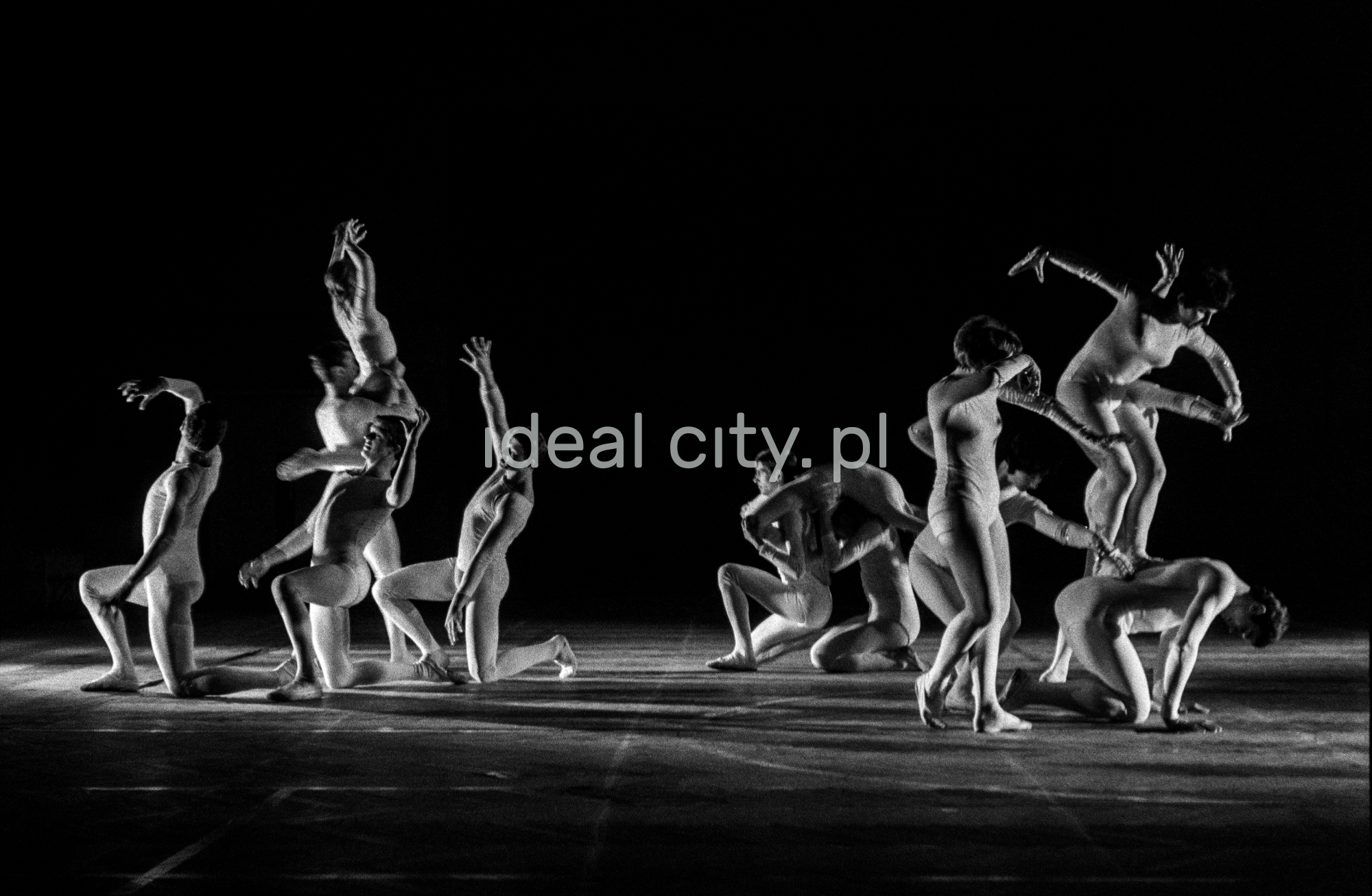 A group of dancers in tight-fitting costumes perform a collective figure on stage with a black background.