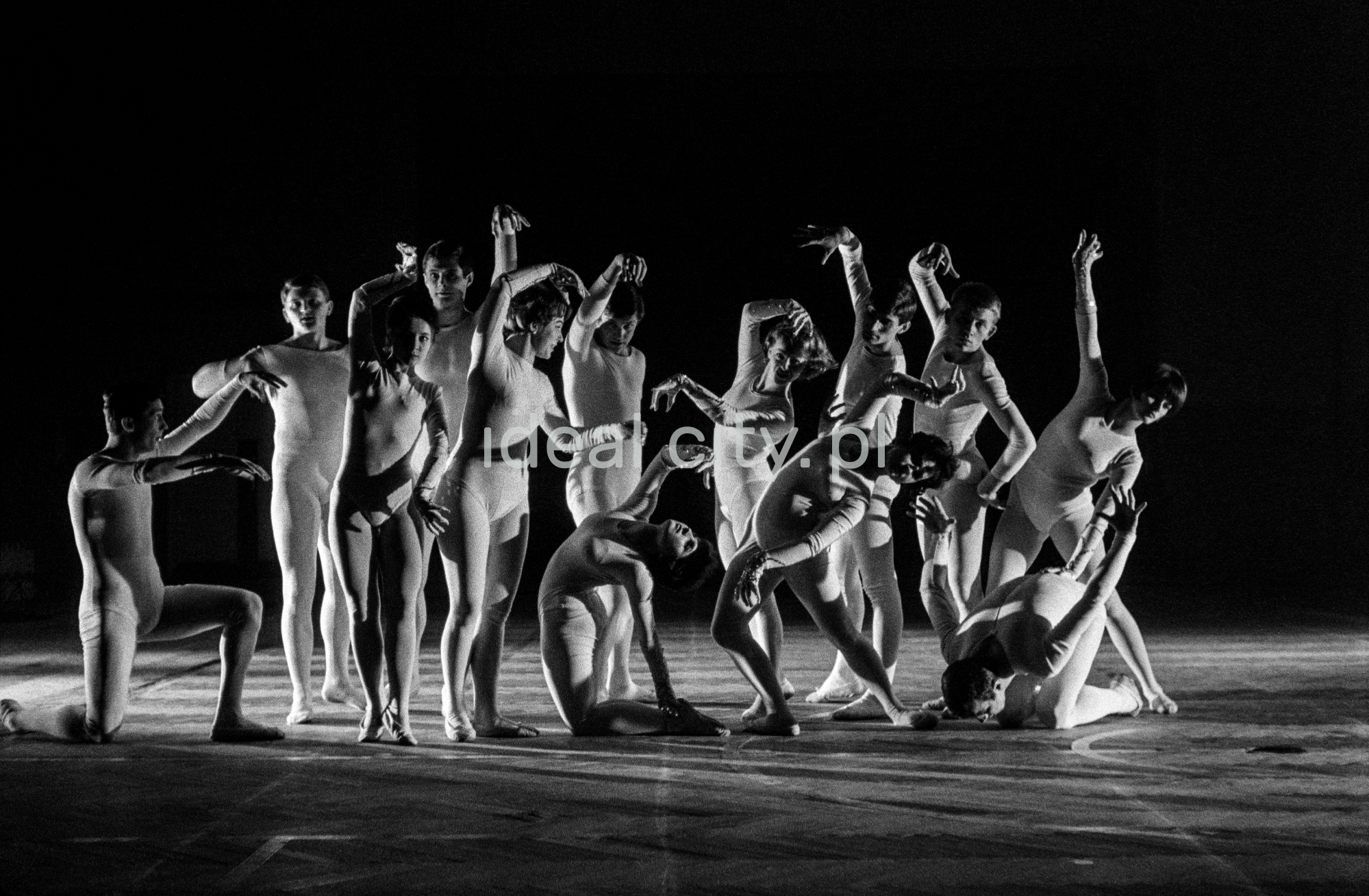 A group of dancers in tight-fitting costumes perform a collective figure on stage with a black background.