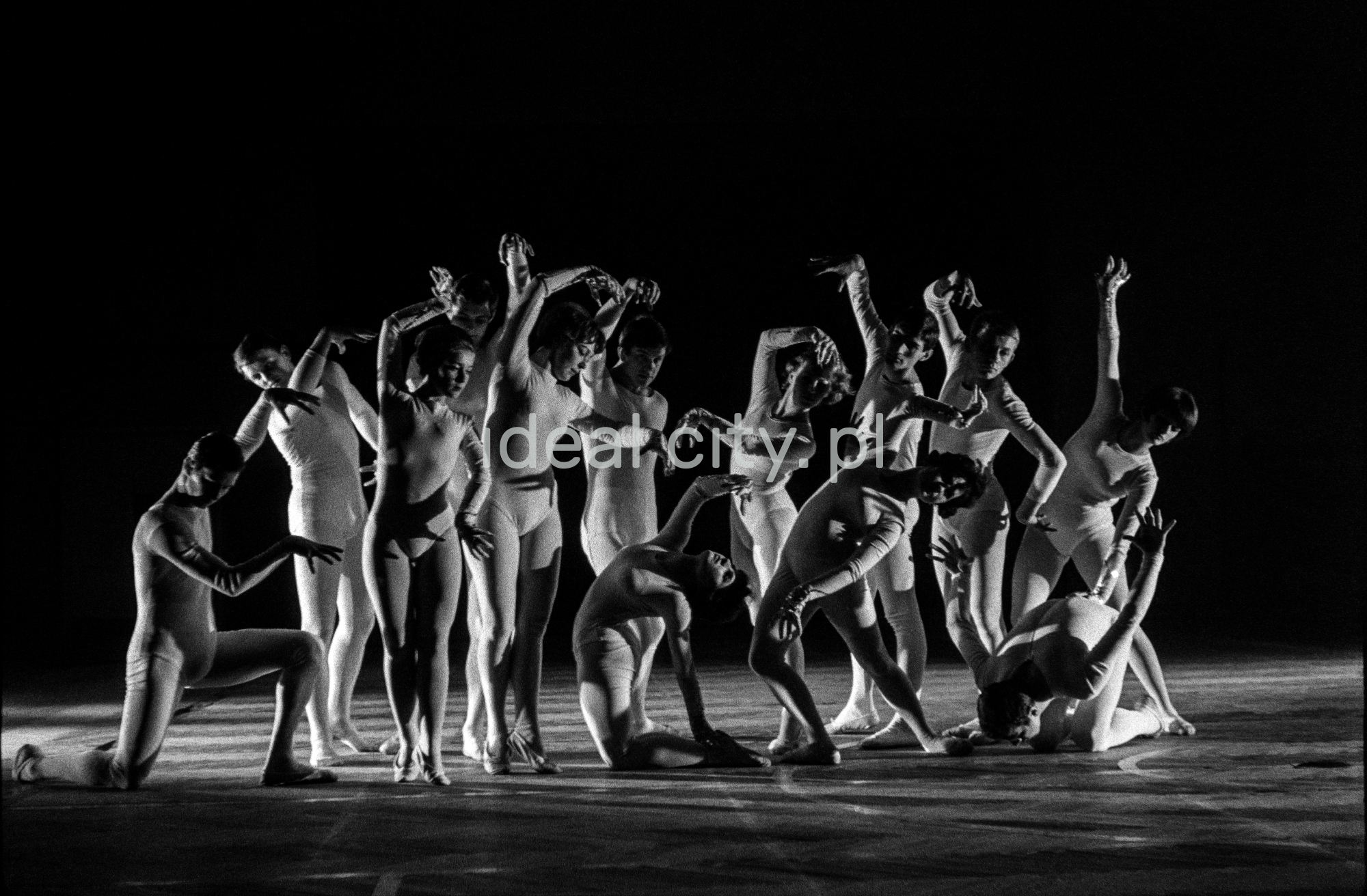A group of dancers in tight-fitting costumes perform a collective figure on stage with a black background.