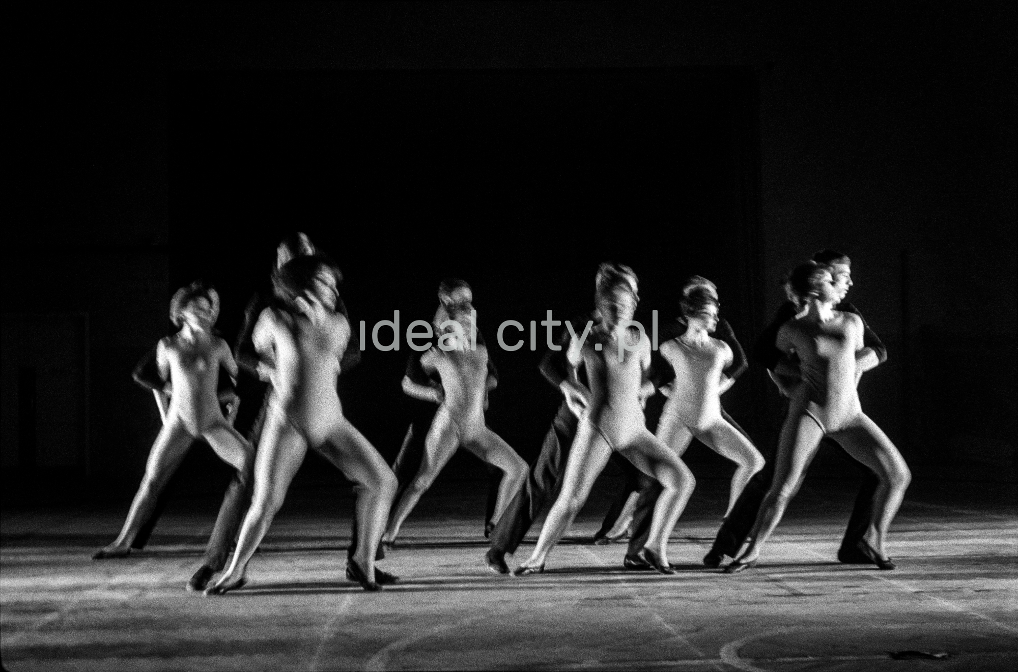 A group of dancers in tight-fitting costumes perform a collective figure on stage with a black background.