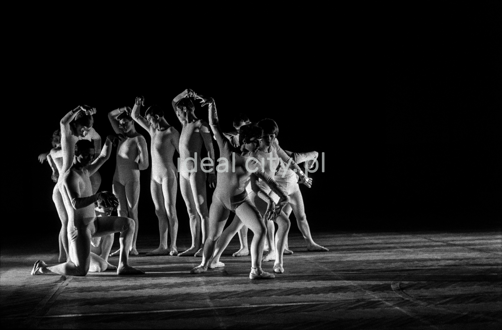 A group of dancers in tight-fitting costumes perform a collective figure on stage with a black background.