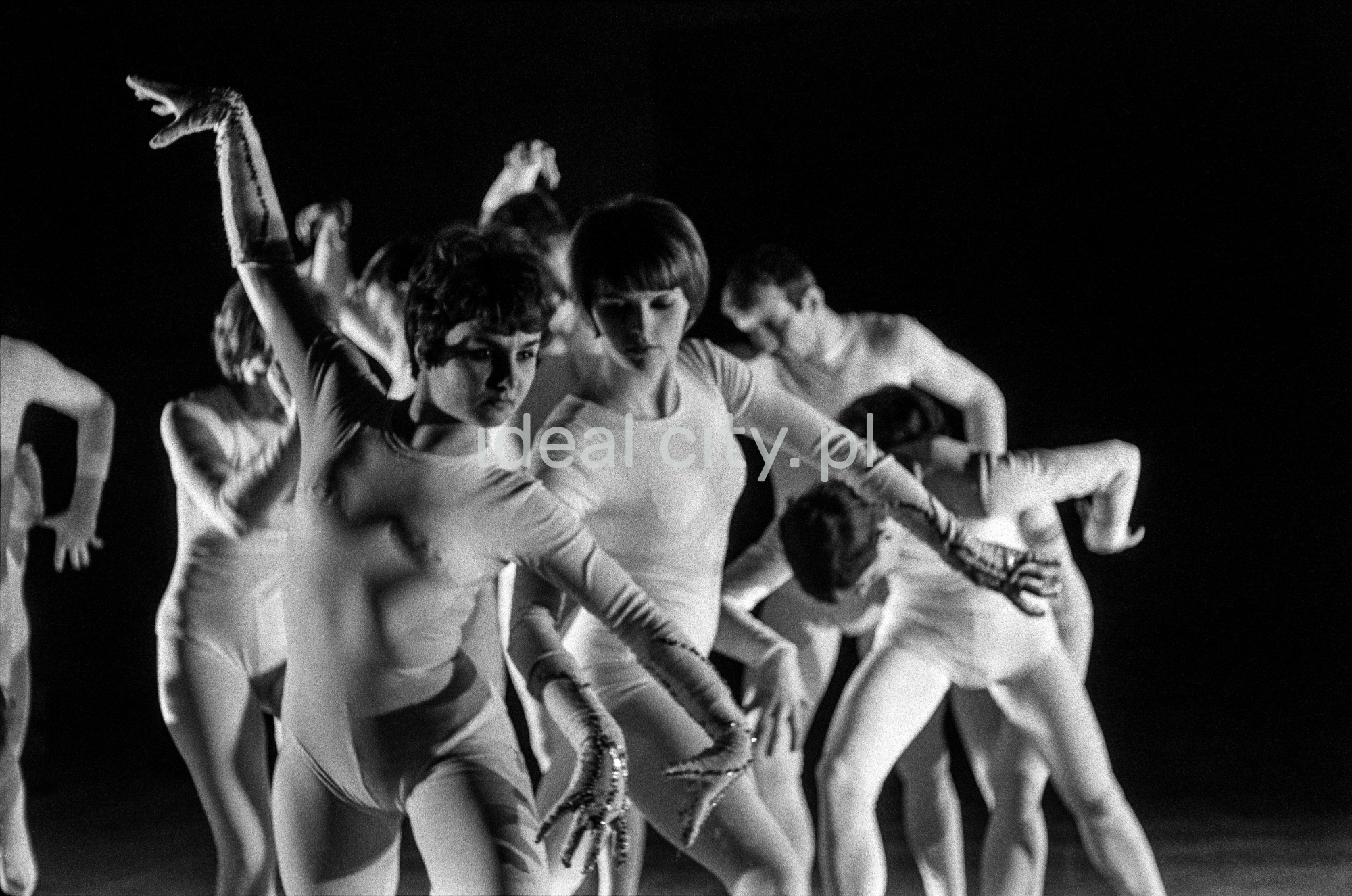 A group of dancers in tight-fitting costumes perform a collective figure on stage with a black background.