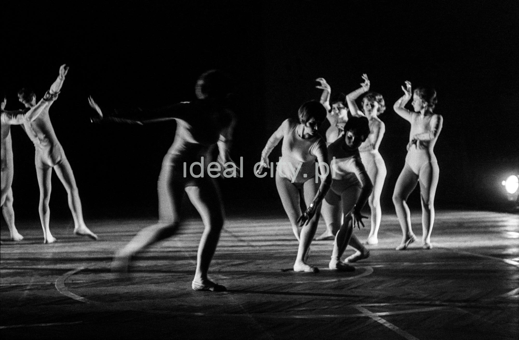 A group of dancers in tight-fitting costumes perform a collective figure on stage with a black background.