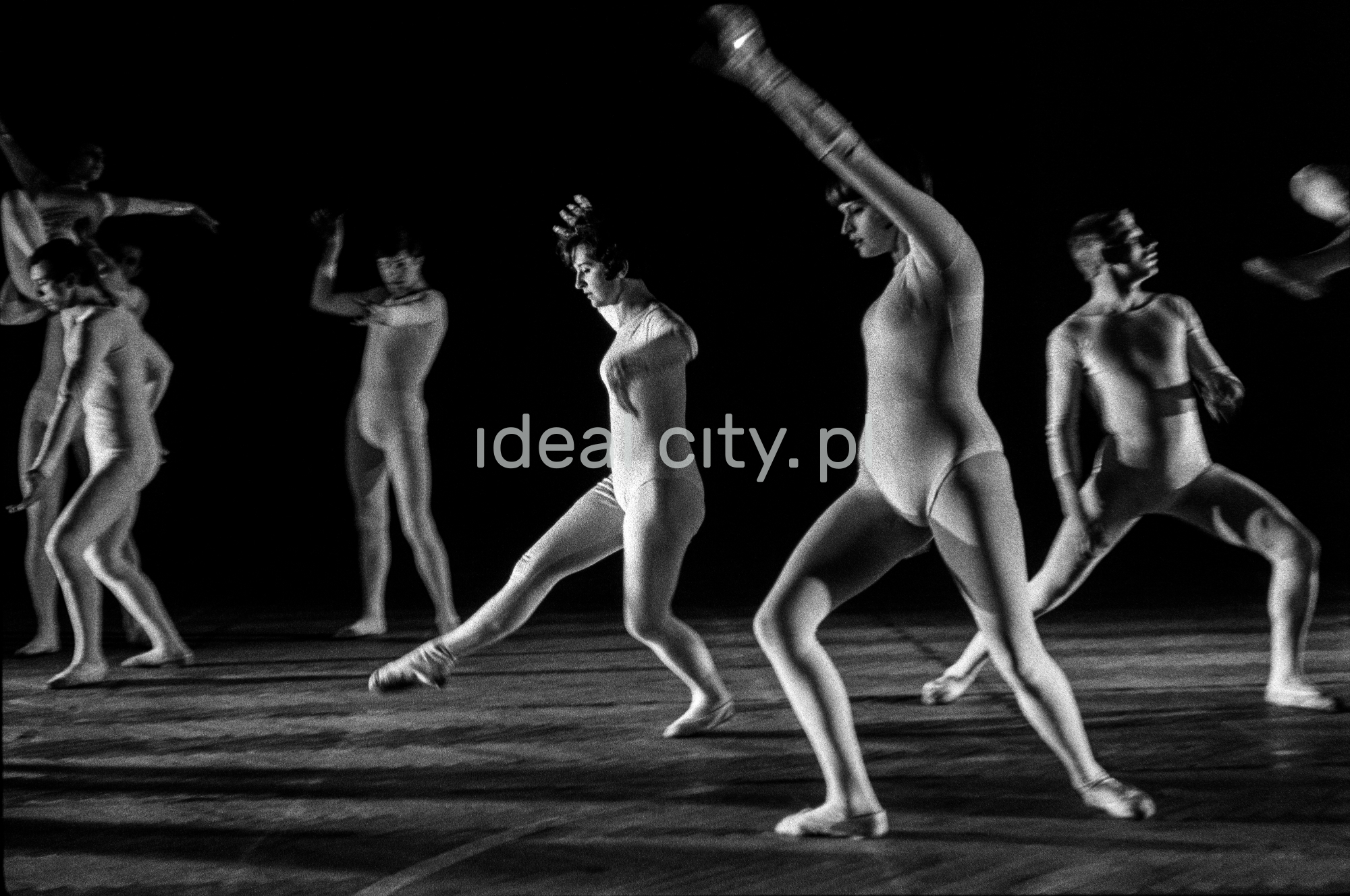 A group of dancers in tight-fitting costumes perform a collective figure on stage with a black background.