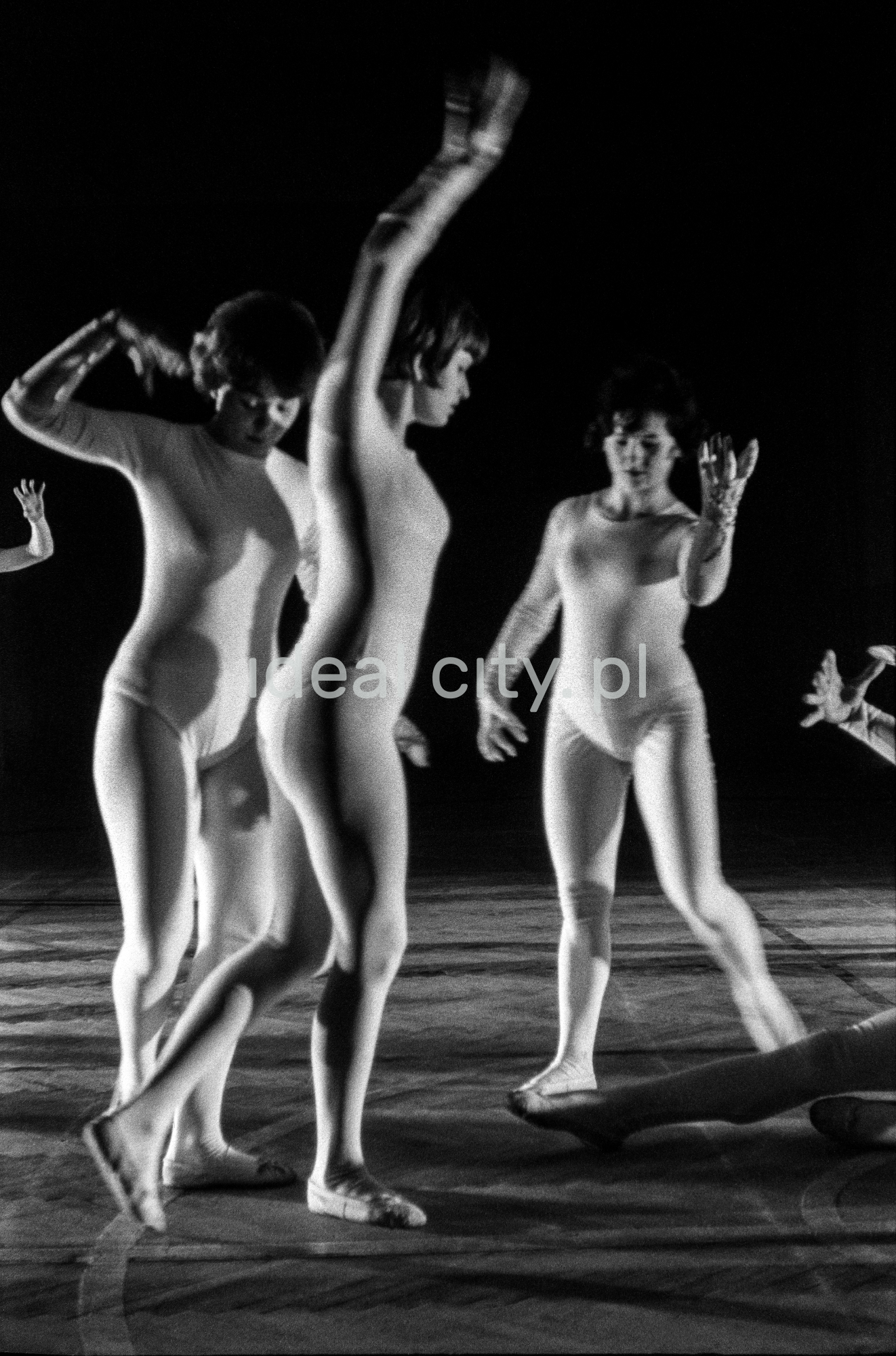 A group of dancers in tight-fitting costumes perform a collective figure on stage with a black background.