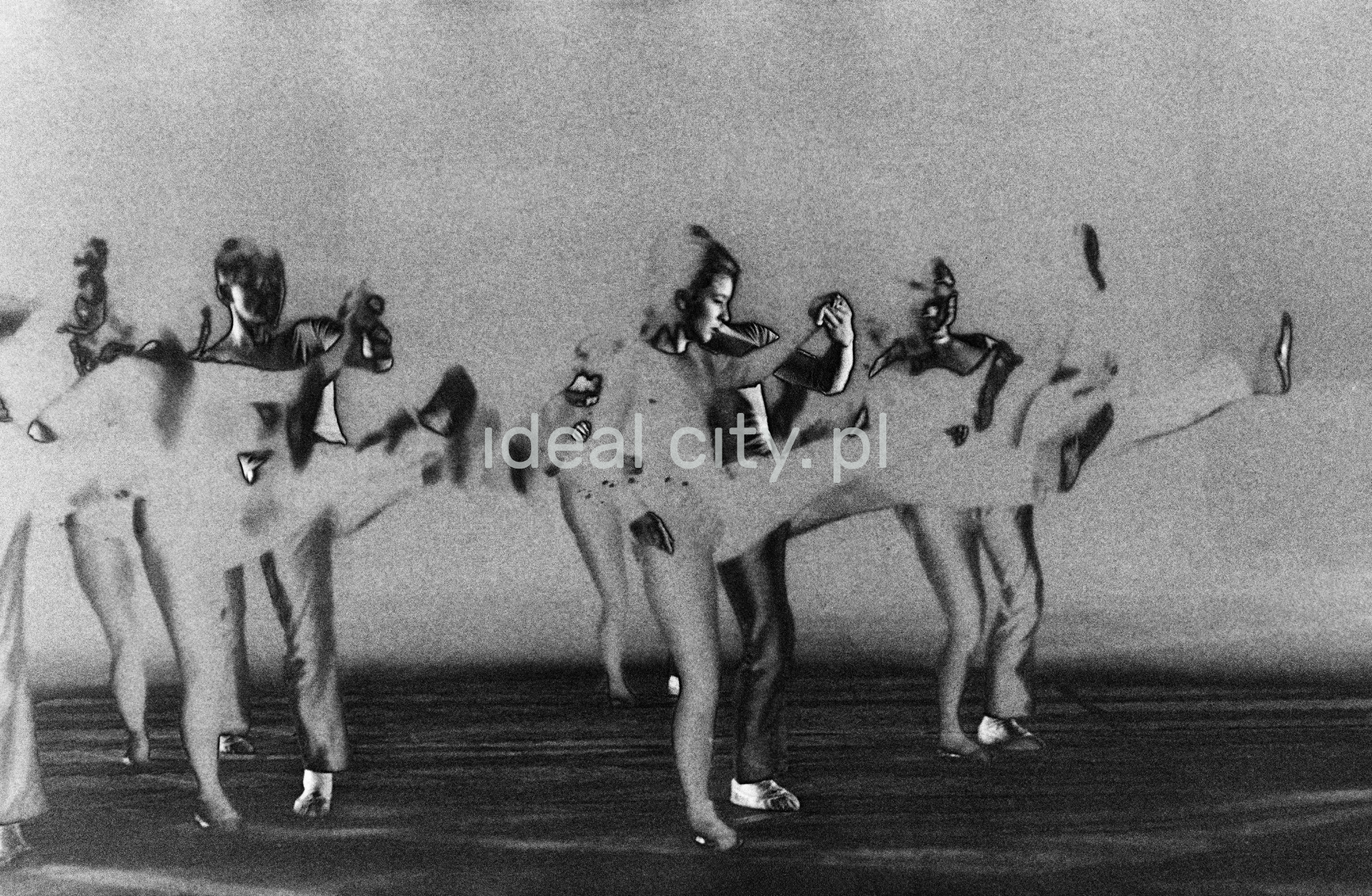 A group of dancers in tight-fitting costumes perform a collective figure on stage with a black background. The negative was solarized.