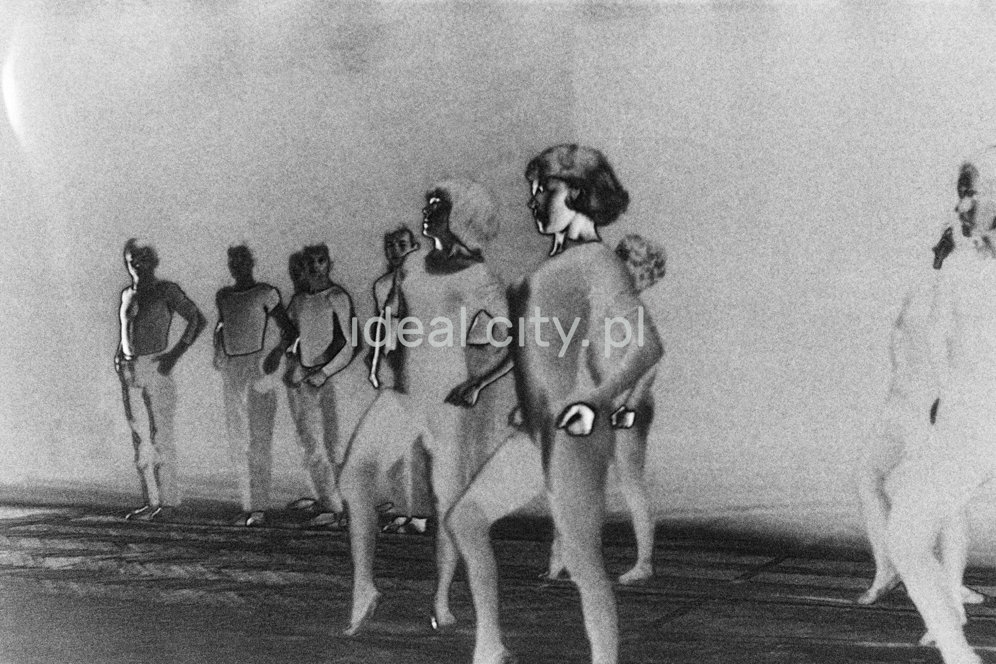 A group of dancers in tight-fitting costumes perform a collective figure on stage with a black background. The negative was solarized.