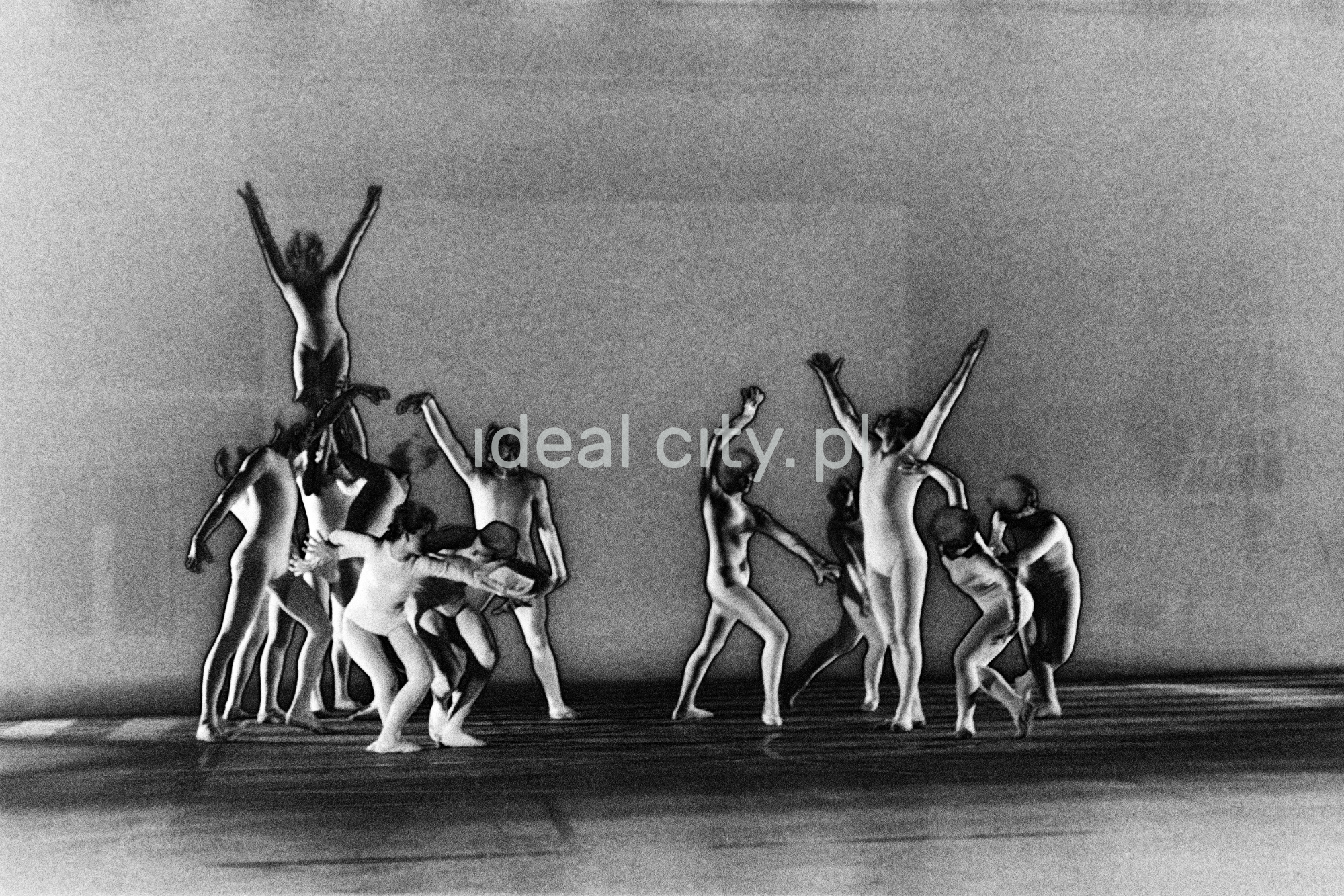 A group of dancers in tight-fitting costumes perform a collective figure on stage with a black background. The negative was solarized.