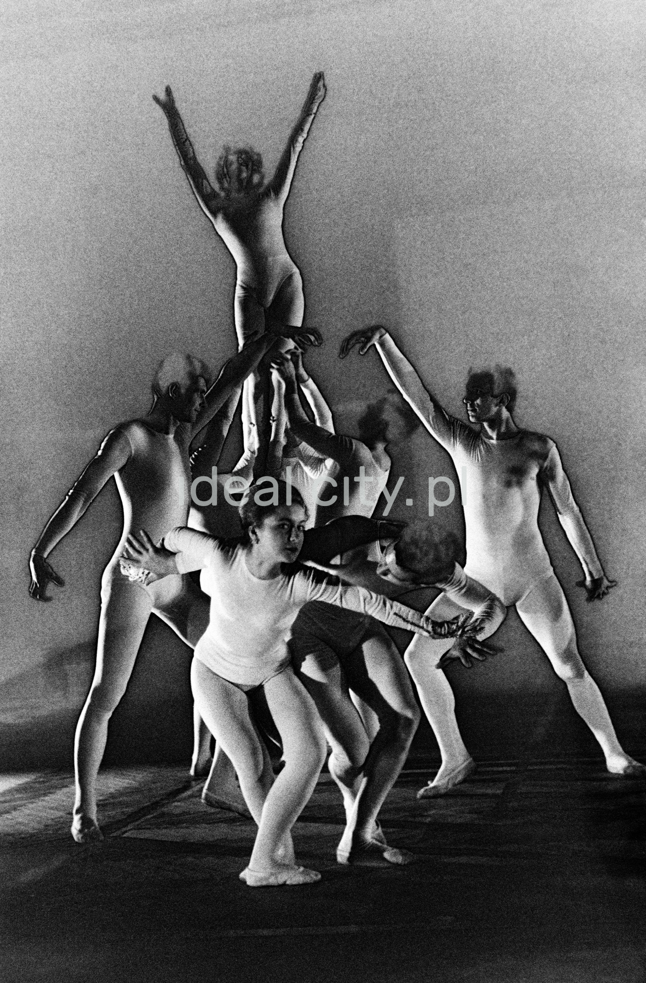 A group of dancers in tight-fitting costumes perform a collective figure on stage with a black background. The negative was solarized.