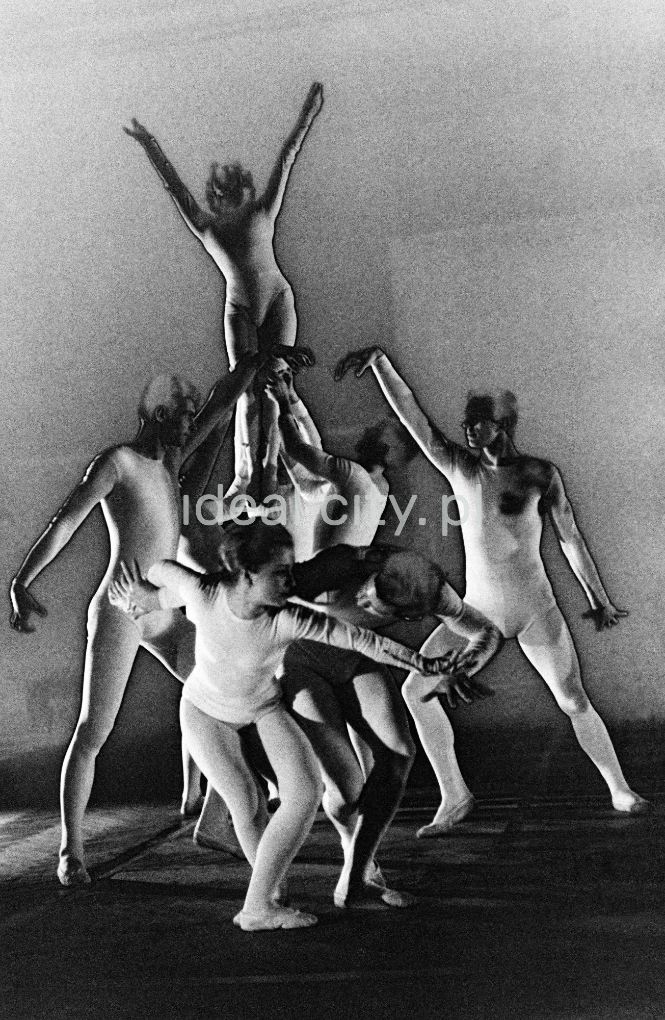 A group of dancers in tight-fitting costumes perform a collective figure on stage with a black background. The negative was solarized.