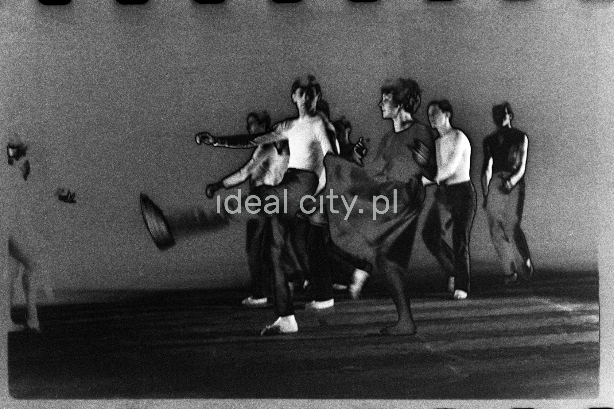 A group of dancers in tight-fitting costumes perform a collective figure on stage with a black background. The negative was solarized.