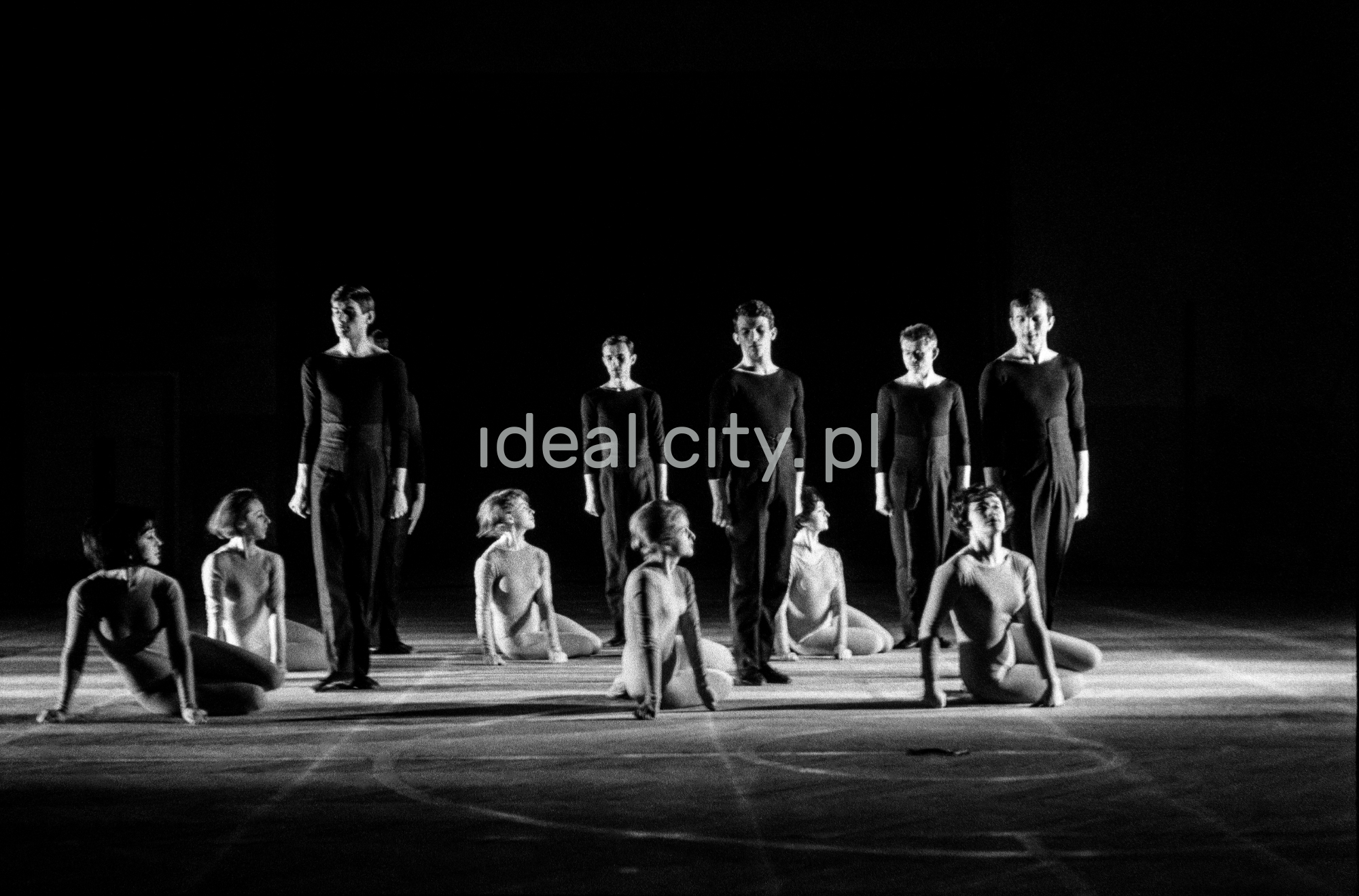 A group of dancers in tight-fitting costumes perform a collective figure on stage with a black background.