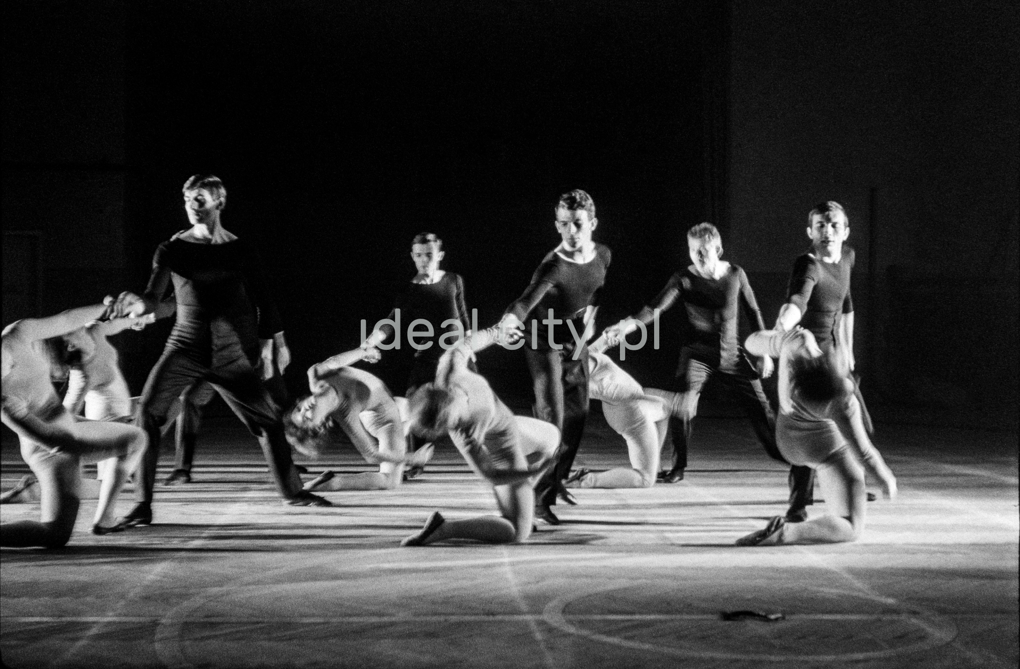 A group of dancers in tight-fitting costumes perform a collective figure on stage with a black background.