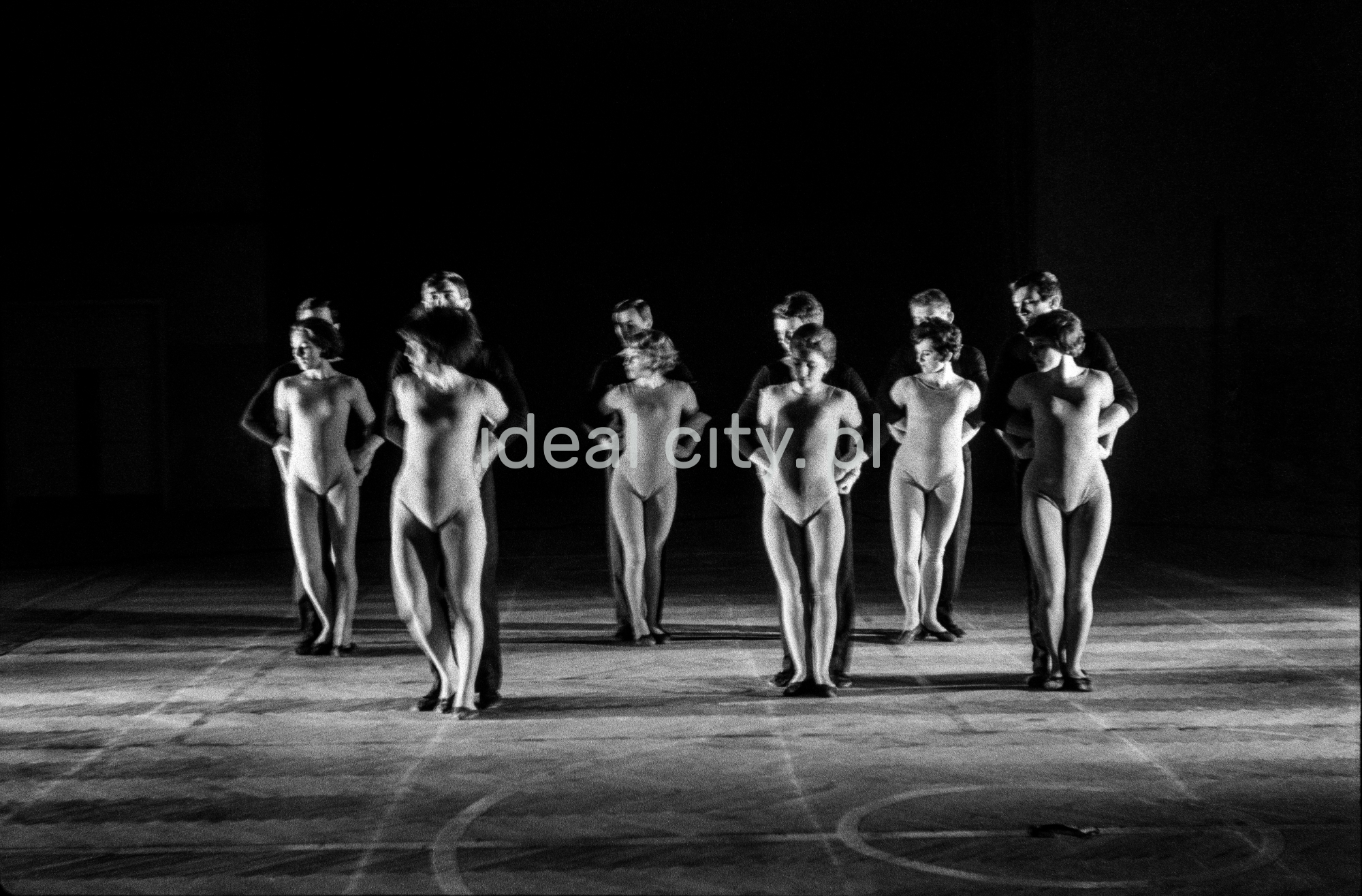 A group of dancers in tight-fitting costumes perform a collective figure on stage with a black background.