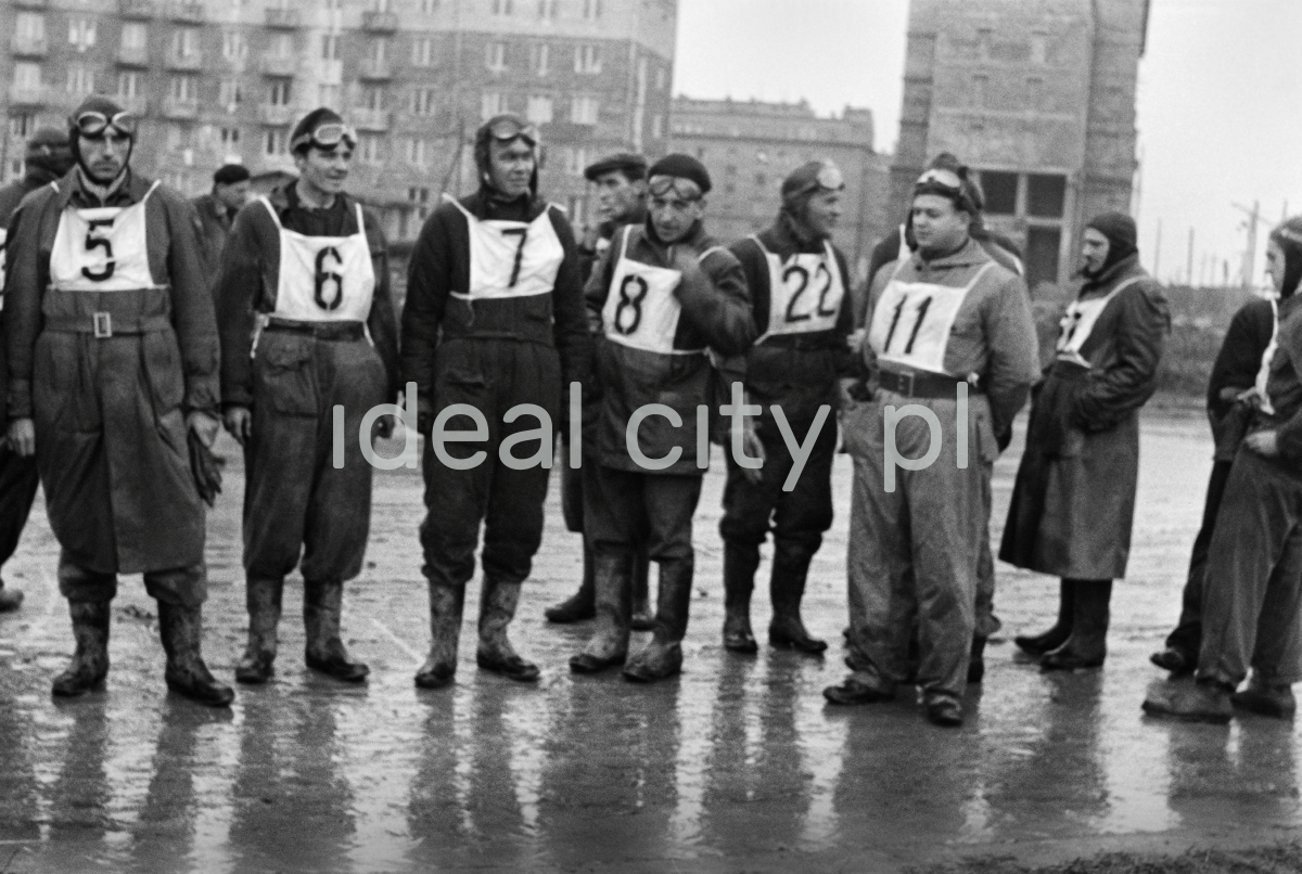 Motorcyclists with numbers on their chests are standing on wet asphalt. Block buildings in the background.