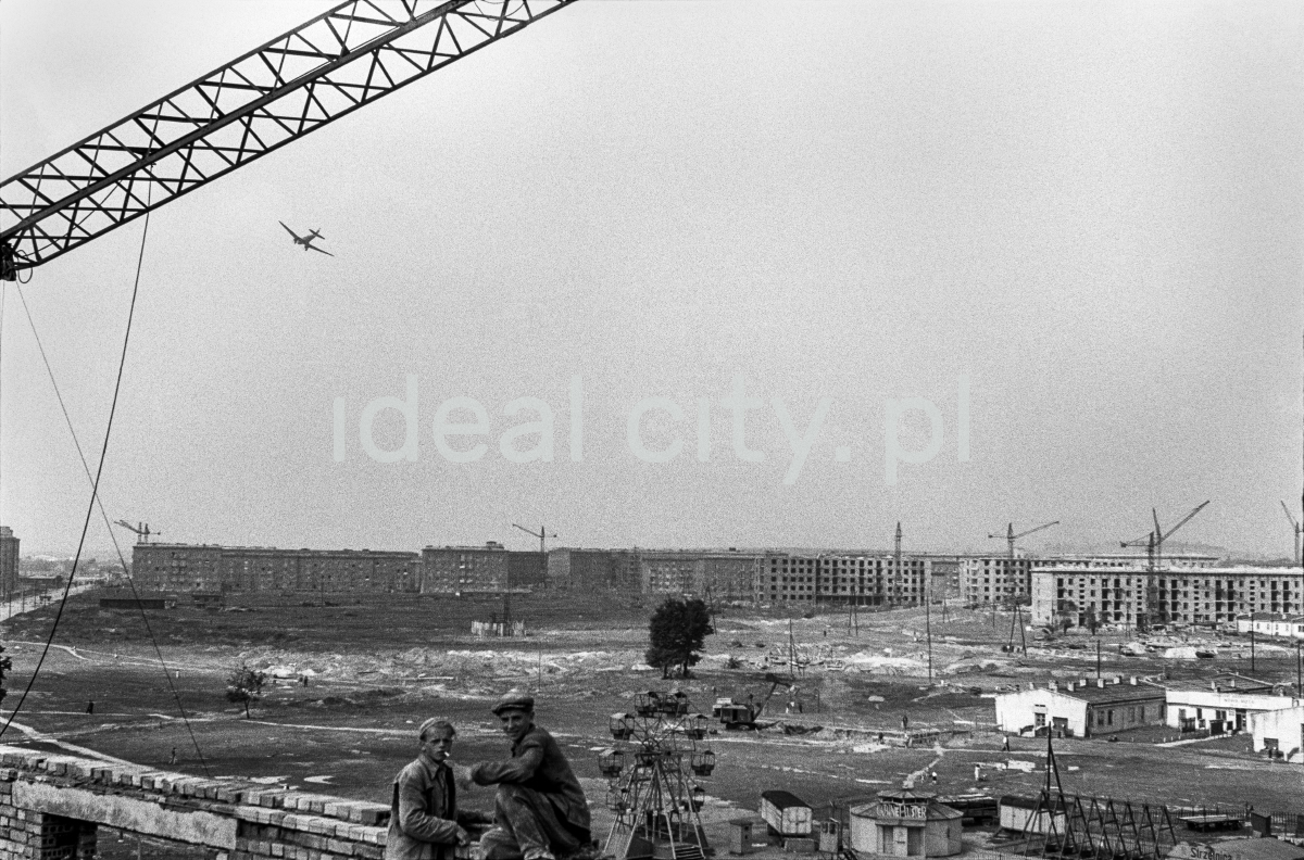 View of the city under construction. In the foreground, there are two small silhouettes of workers, a plane is flying in the sky.