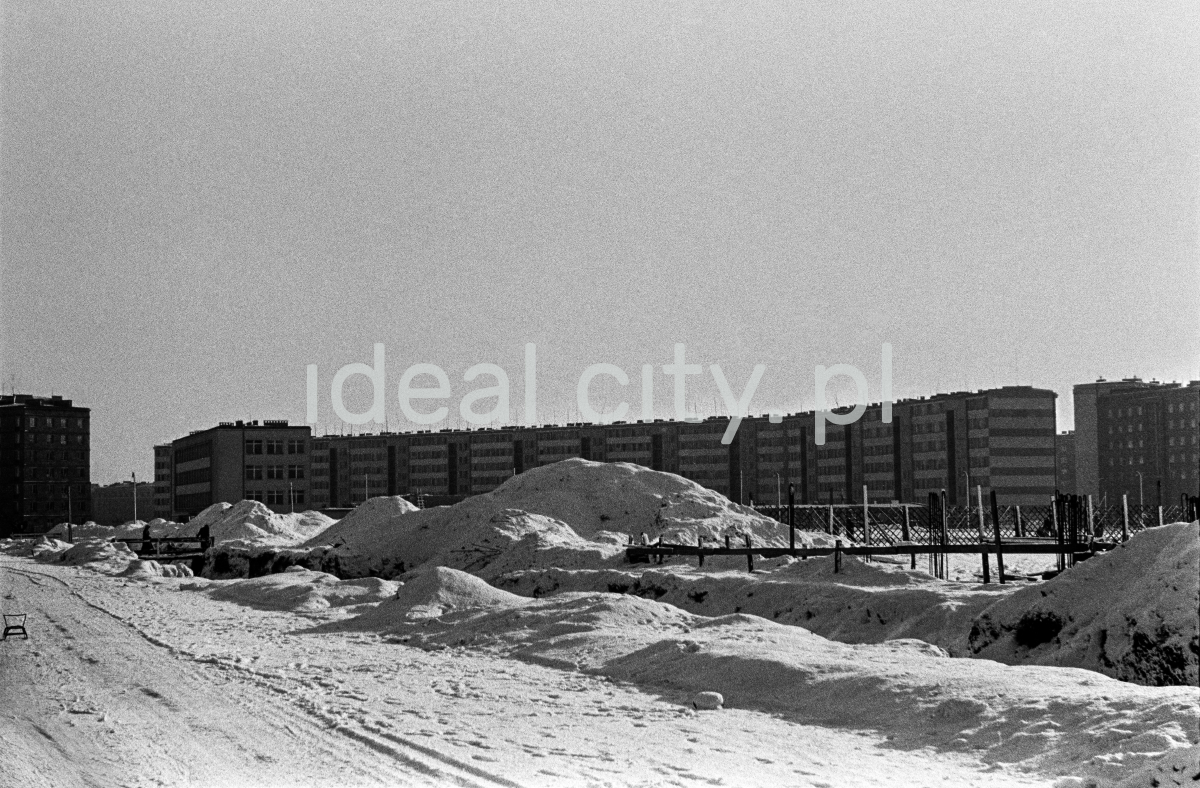 A view of a snow-covered square in the background of a low, wide apartment block.