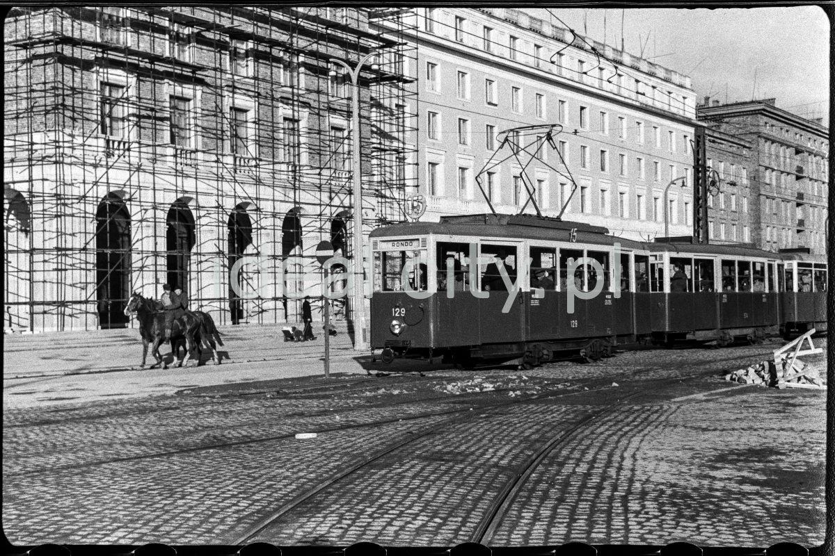 A tram passes in front of a monumental residential building covered with scaffolding, two men are riding on the paving next to it.