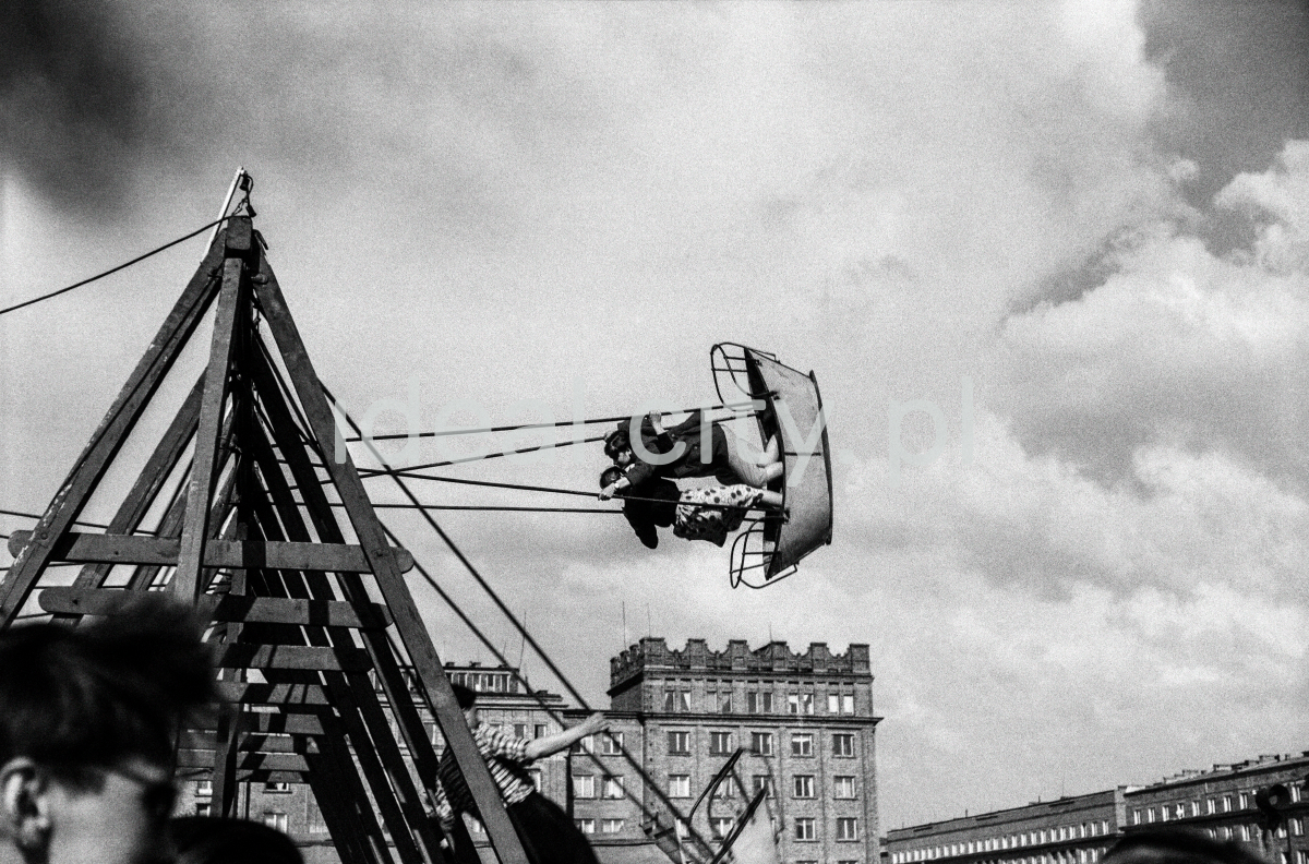 Two girls lean out to their fullest on a boat-shaped swing, monumental residential buildings in the background.