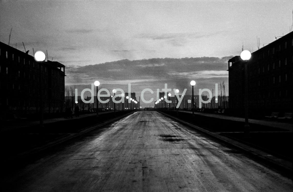 A view at dawn on a wide avenue lit by street lamps, blocks on both sides.