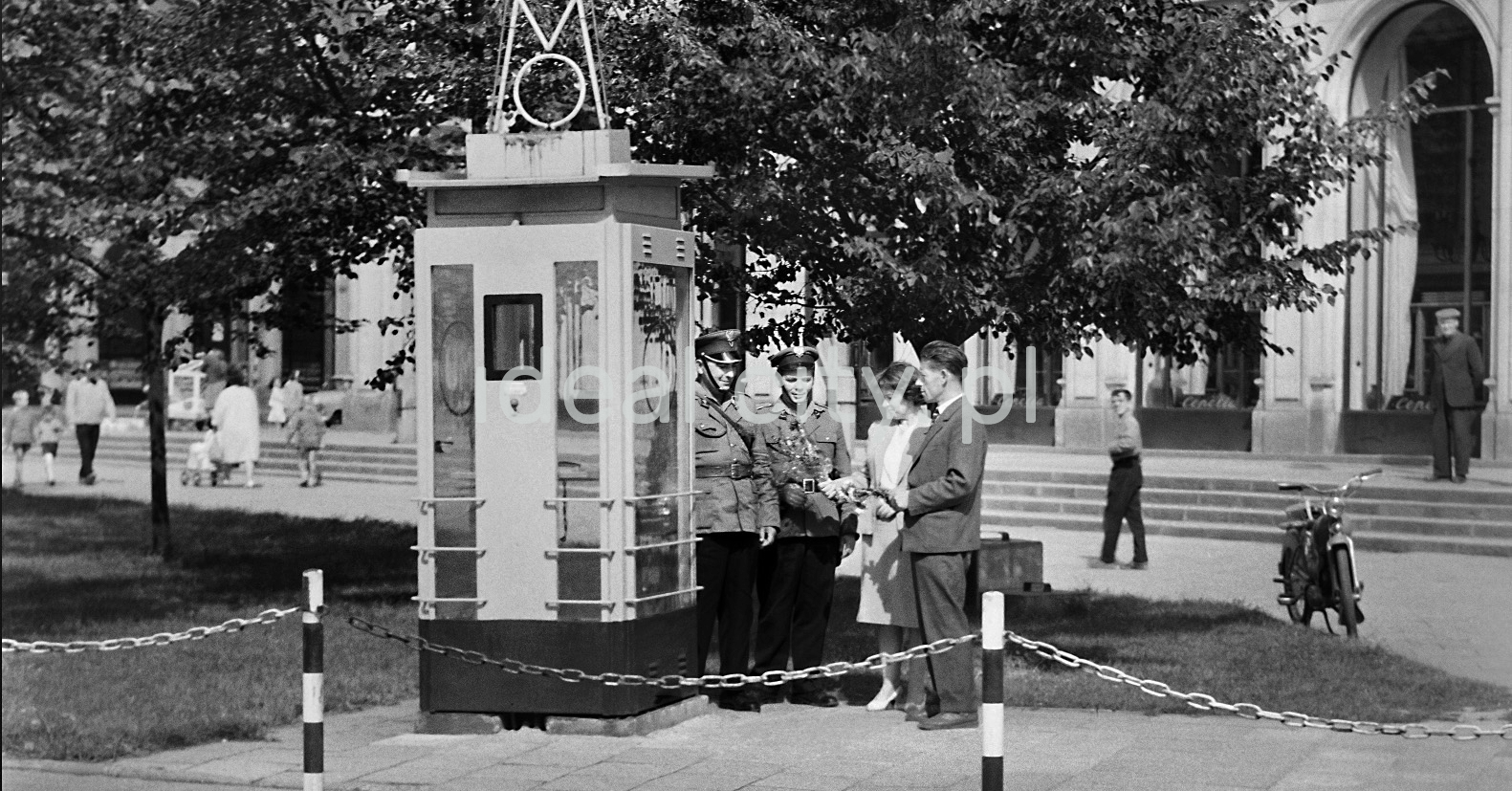 A young woman and a man in festive costumes hand over carnations to a policeman who is leaning towards them from the booth.