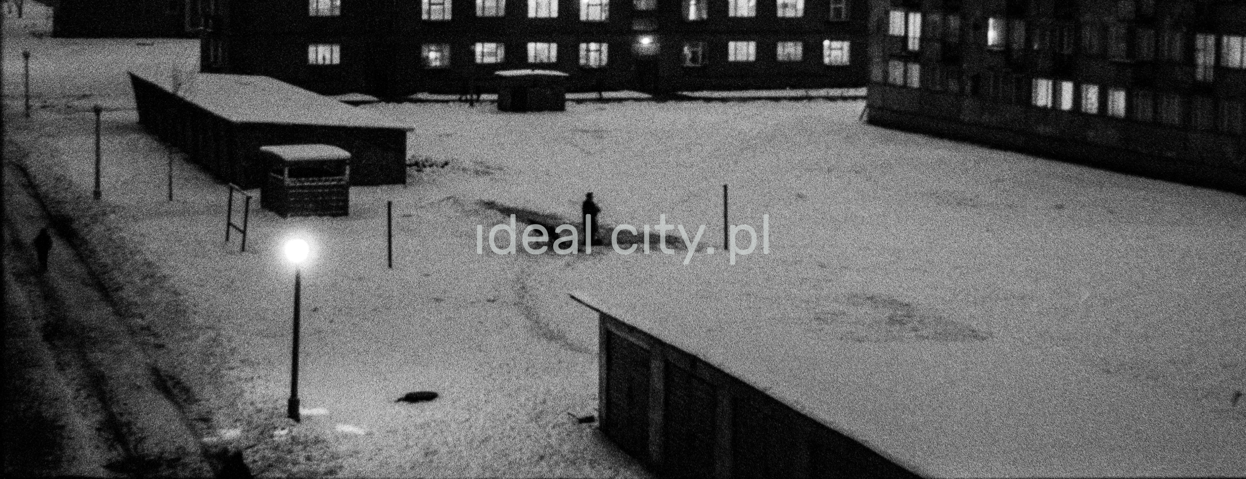 A night view from the window of the snow-covered yard between the blocks, a figure in a coat in the center.
