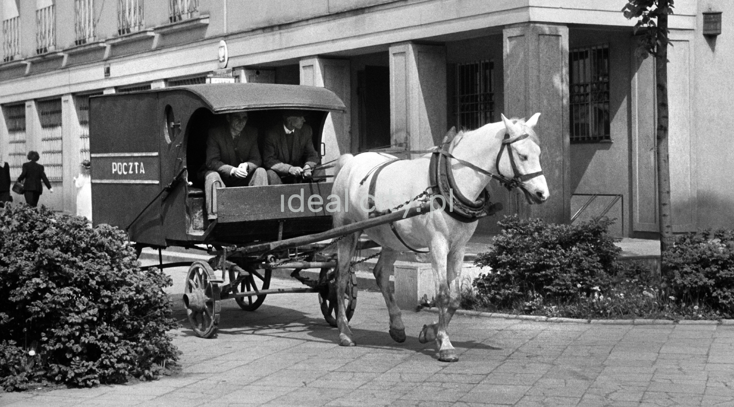 A horse-drawn cart with the inscription "post", drawn by a white horse, rides along the sidewalk next to the building.