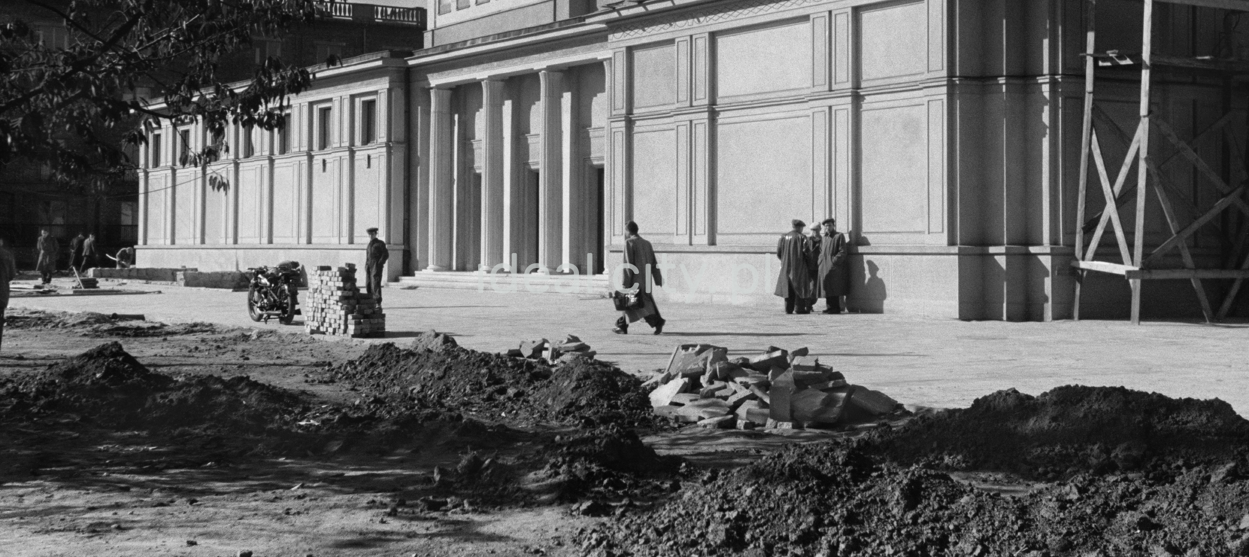 Several men in coats are standing by the wall of the monumental building, more are walking in front of it, bricks in the foreground.
