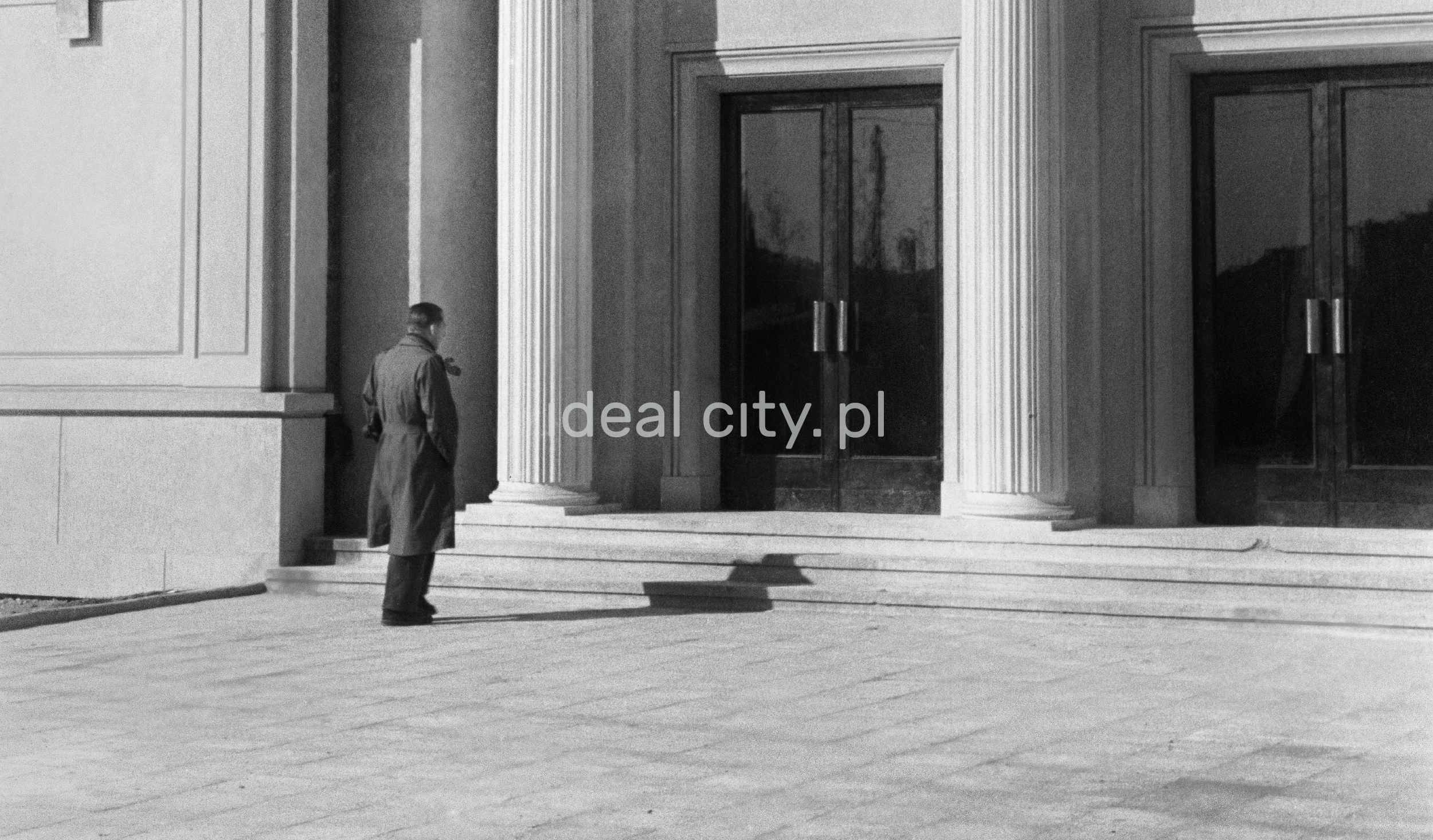 The man in the coat stands in front of the monumental entrance to the building.