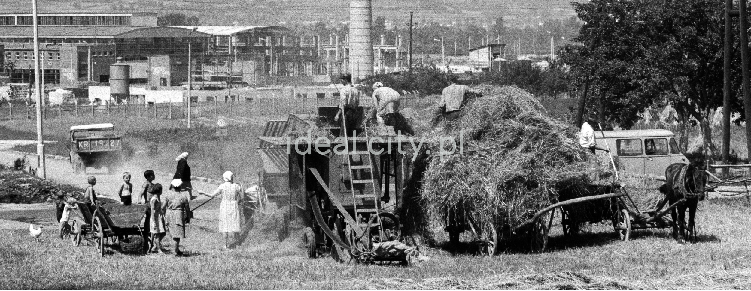 The family uses a simple harvester to collect hay from the meadow onto a cart drawn by a horse. In the background, a chimney and factory buildings.