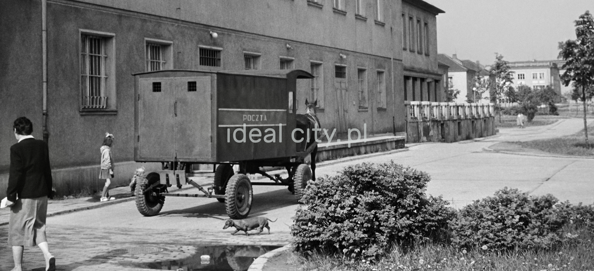 A horse-drawn postal cart goes along the building., passers around.