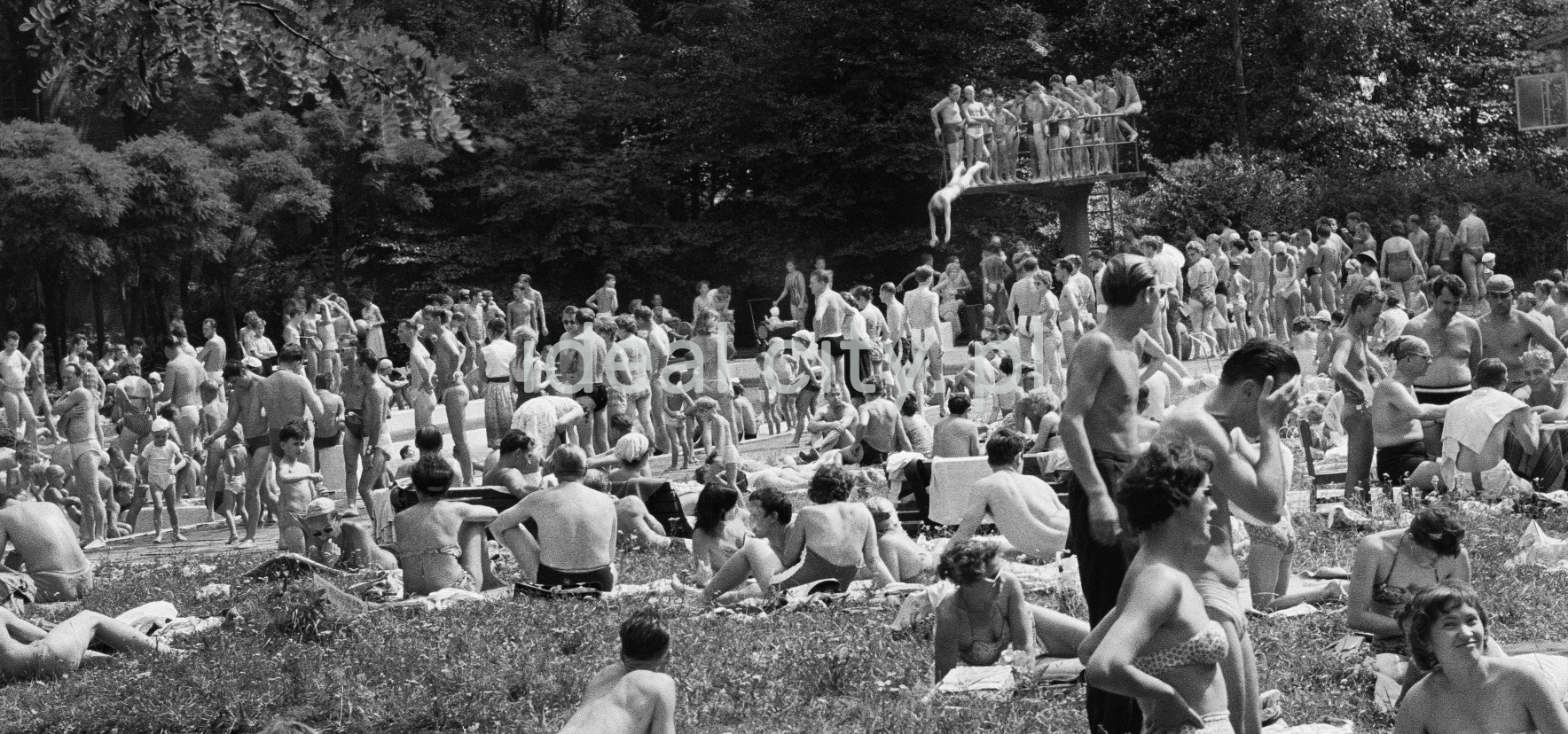 A crowd of people relaxing by the pool, in the background someone jumps into the water from a low springboard.
