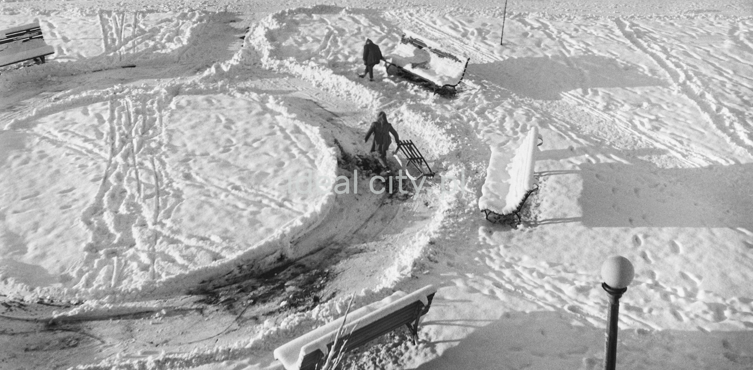 A shot of a snow-covered pavement where children are dragging a sledge.
