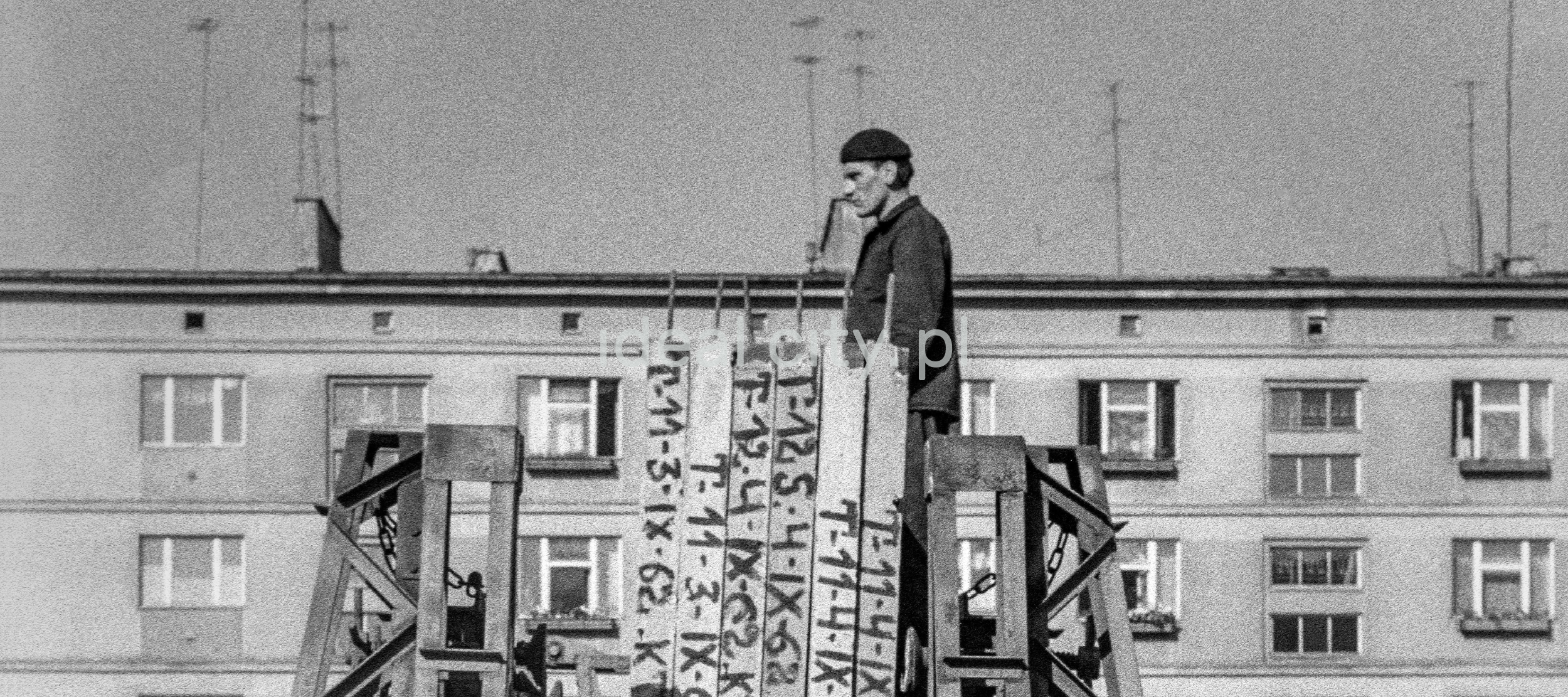 A man in work clothes and a beret protrudes in a monumental manner above the prefabricated panels gathered in front of the apartment block.