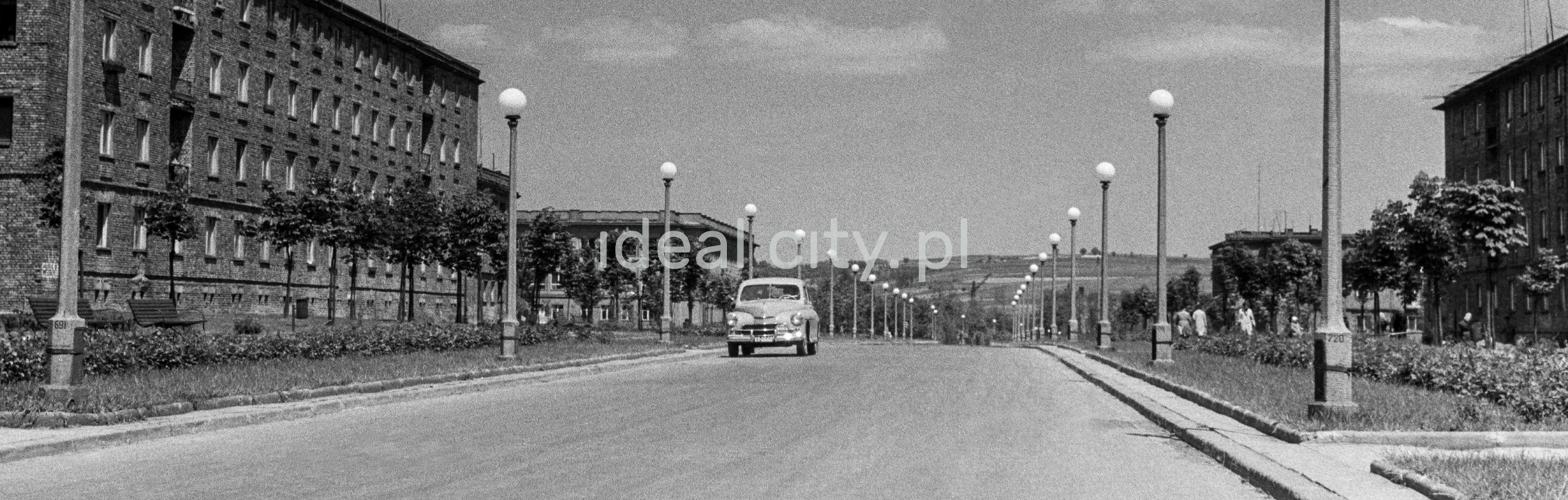 The perspective of a street along which there are two rows of street lamps and apartment blocks, a car parked on the road.
