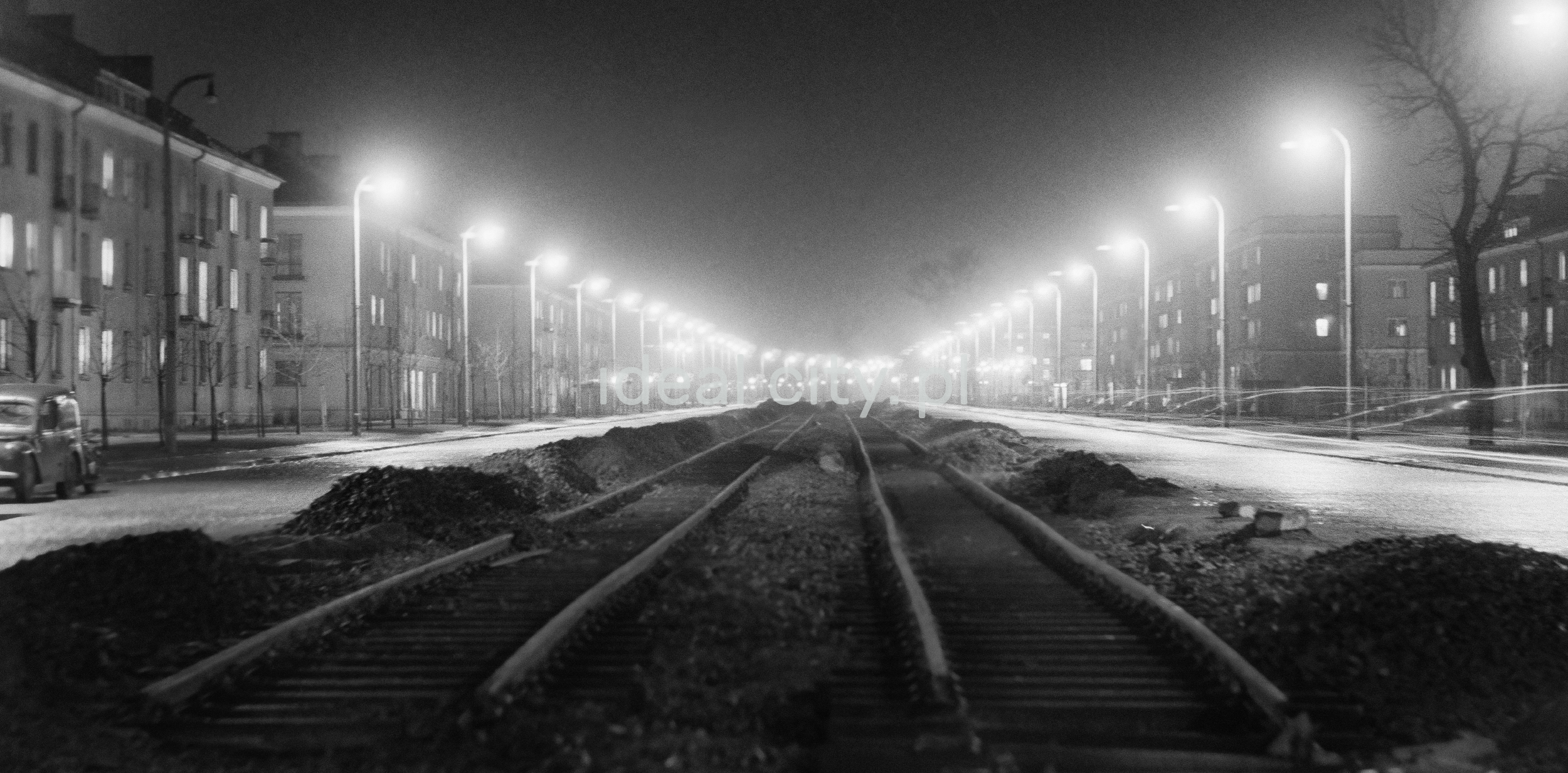 A night perspective of the street, photographed from the center of tram tracks, with apartment blocks on the sides.