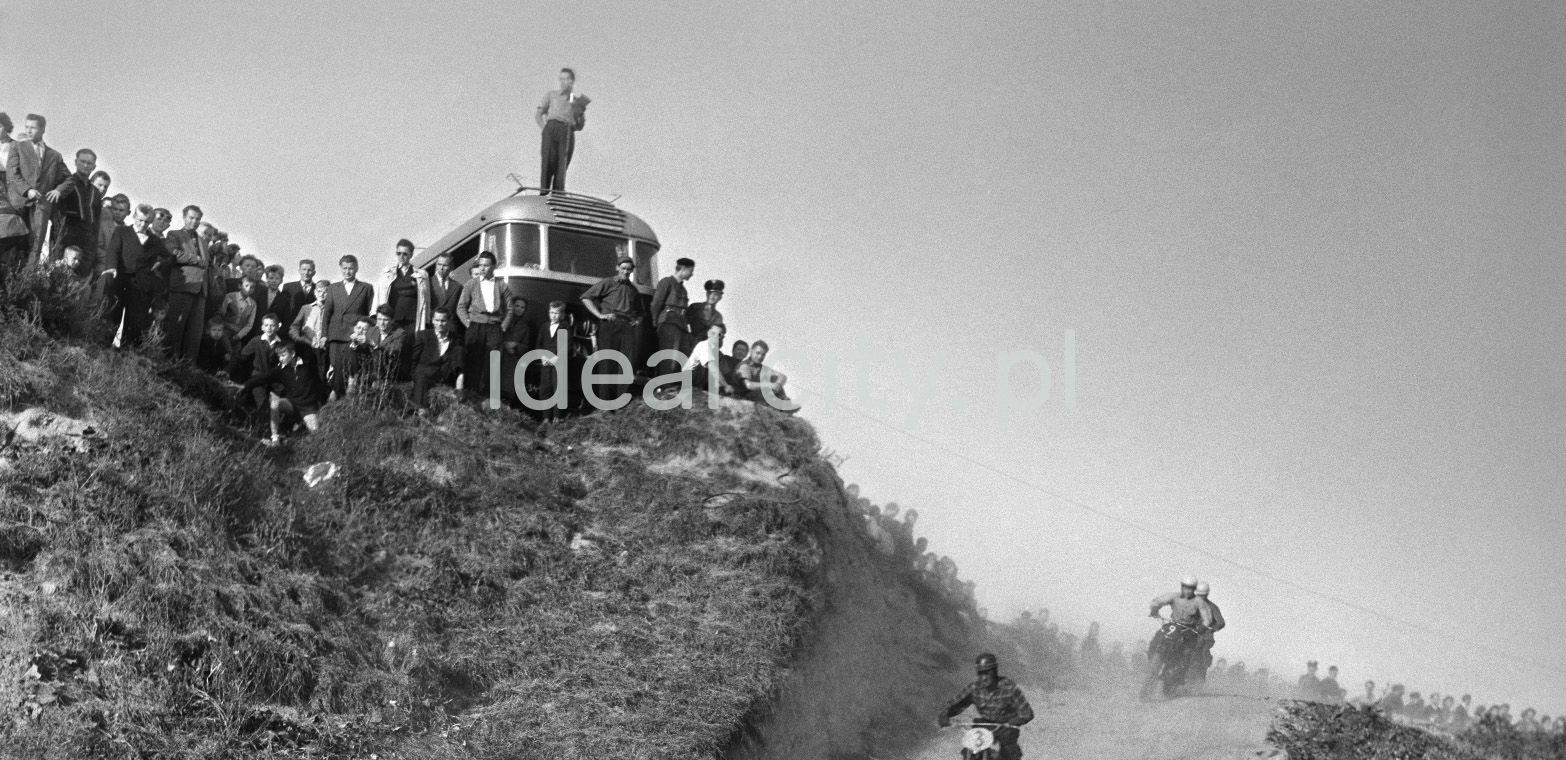Motorcyclists rush down the gravel road, onlookers rush above.